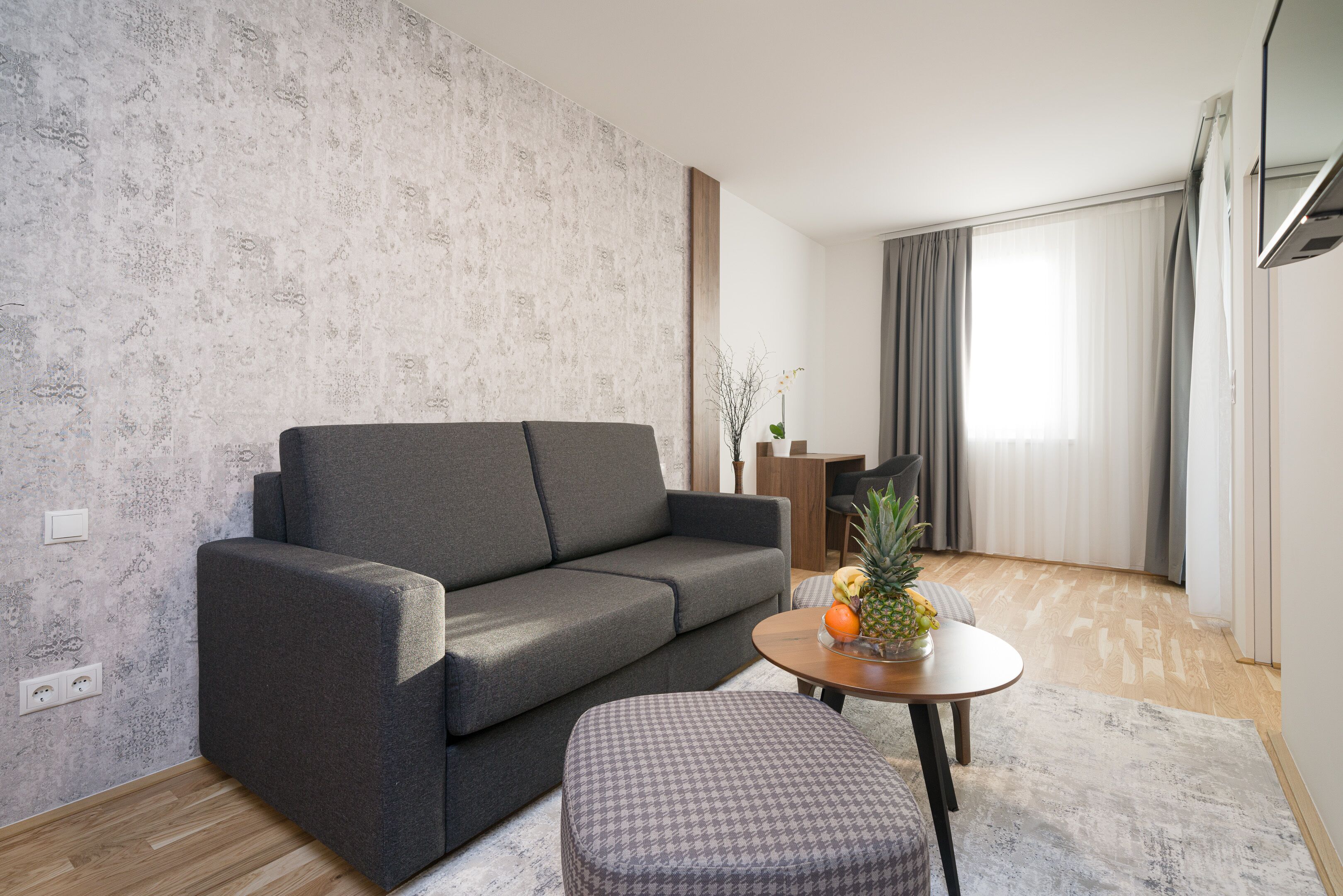 A modern living room with a grey sofa, a small round table with fruits, and a patterned ottoman.