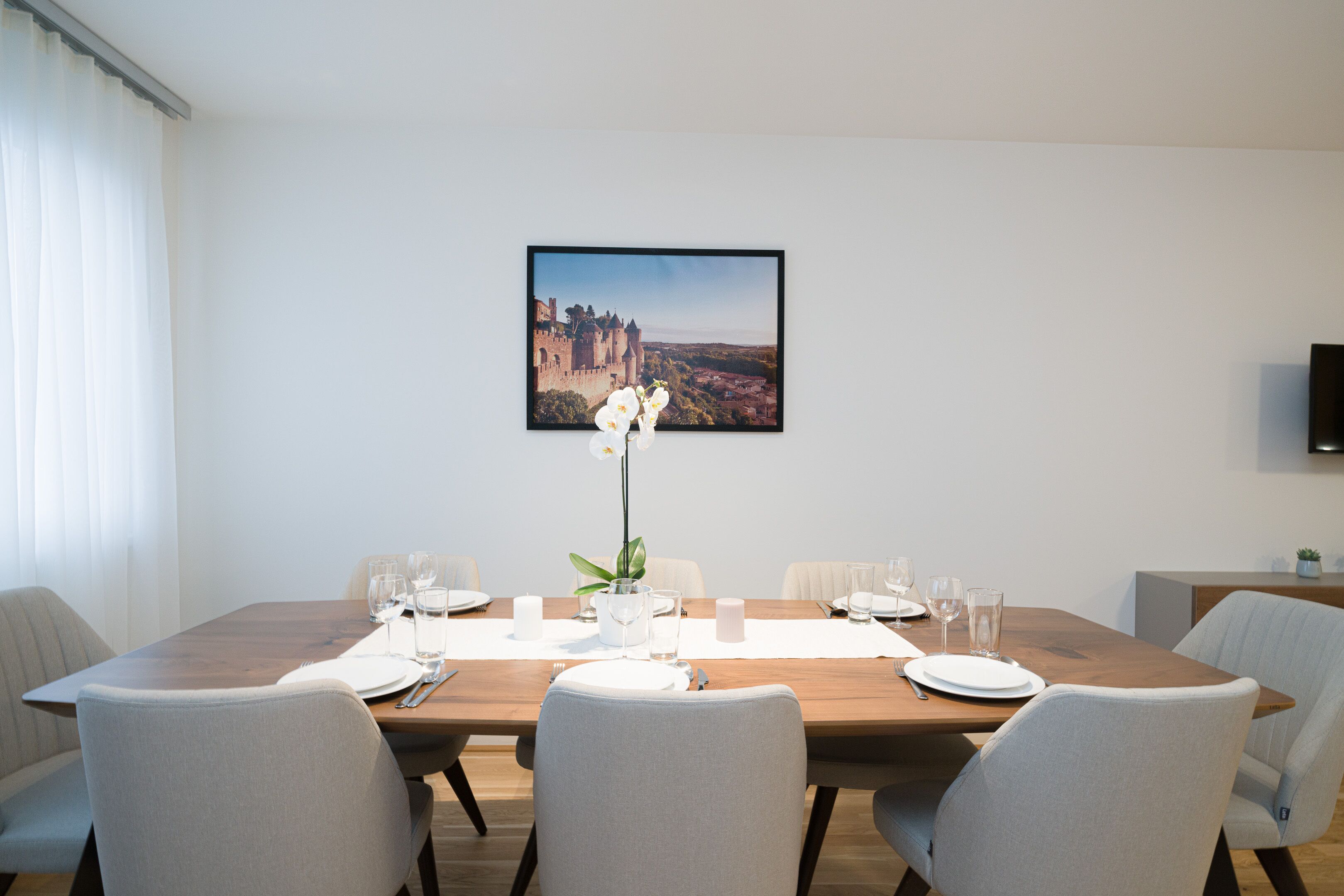 Een eetruimte met een houten tafel voor zes personen, witte stoelen, een lijst met een afbeelding van een kasteel en een potorkidee.