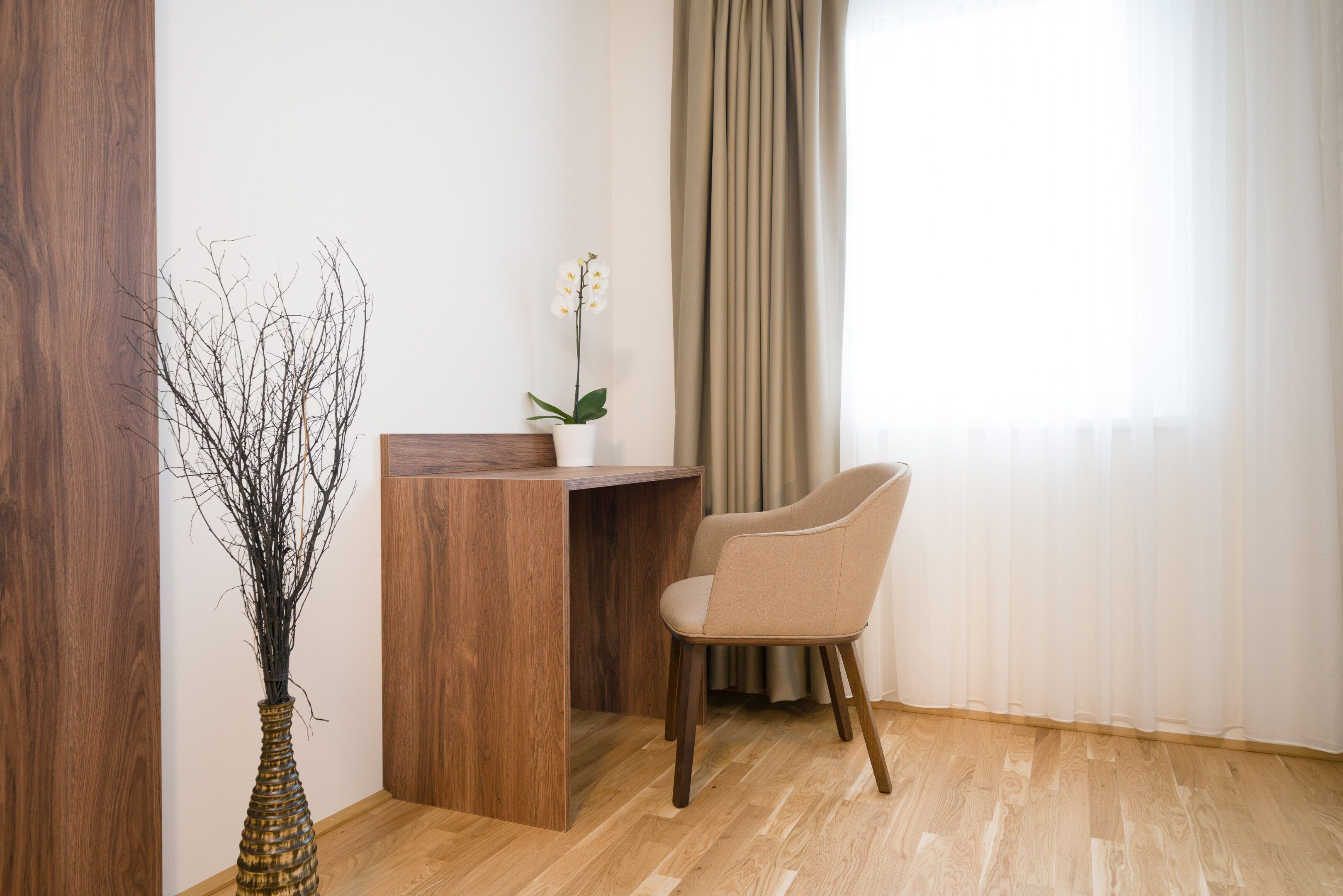 Quarto com escrivaninha de madeira, cadeira bege, um vaso com galhos e janela com cortinas.