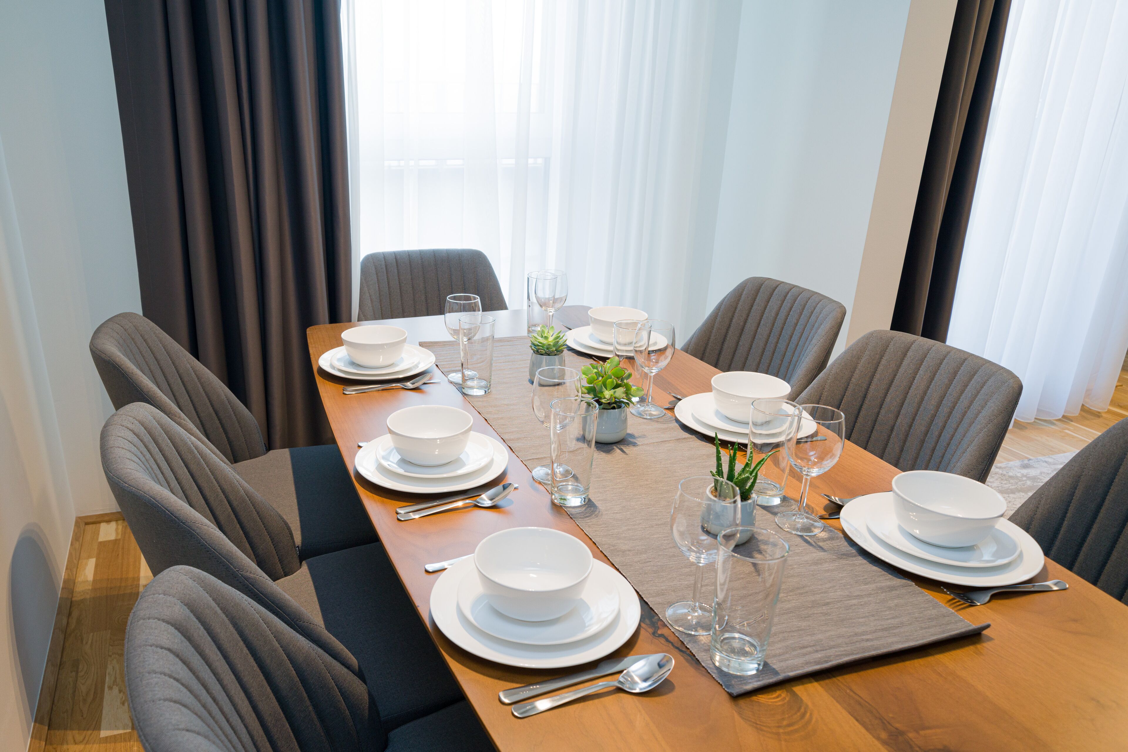 A dining table set for six with white plates, bowls, and glasses, surrounded by grey upholstered chairs.
