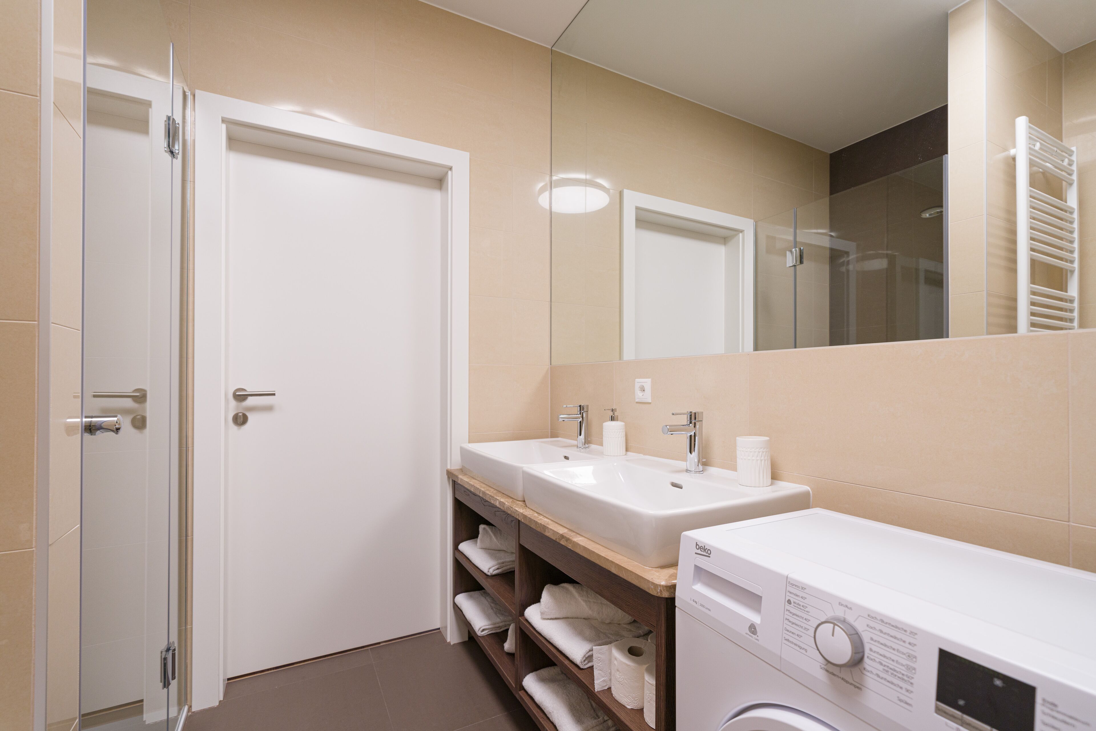 A modern bathroom with two sinks, a mirror, a shower, and a washing machine.