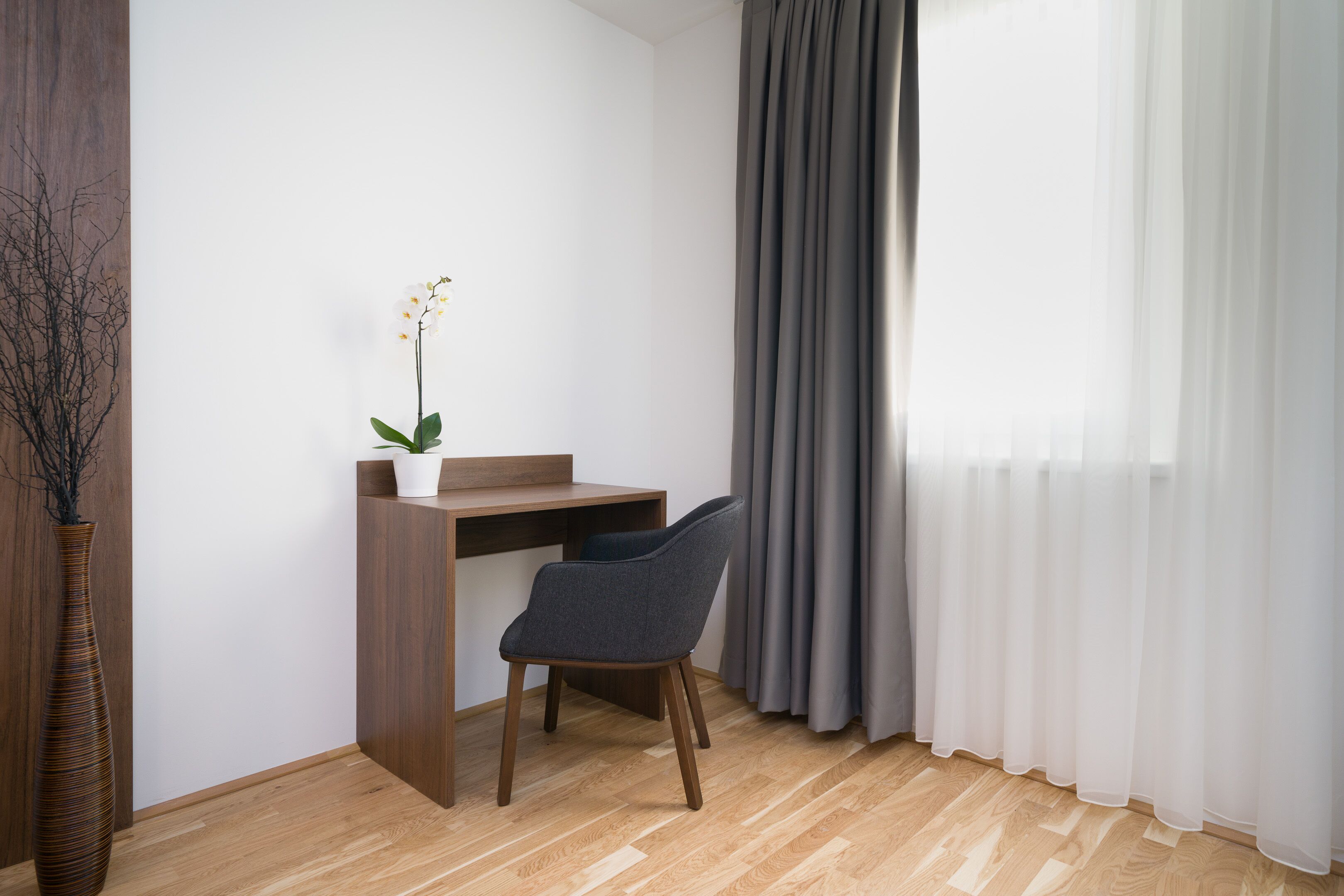A room with a wooden desk, a chair, a potted plant, and a large window with sheer curtains.