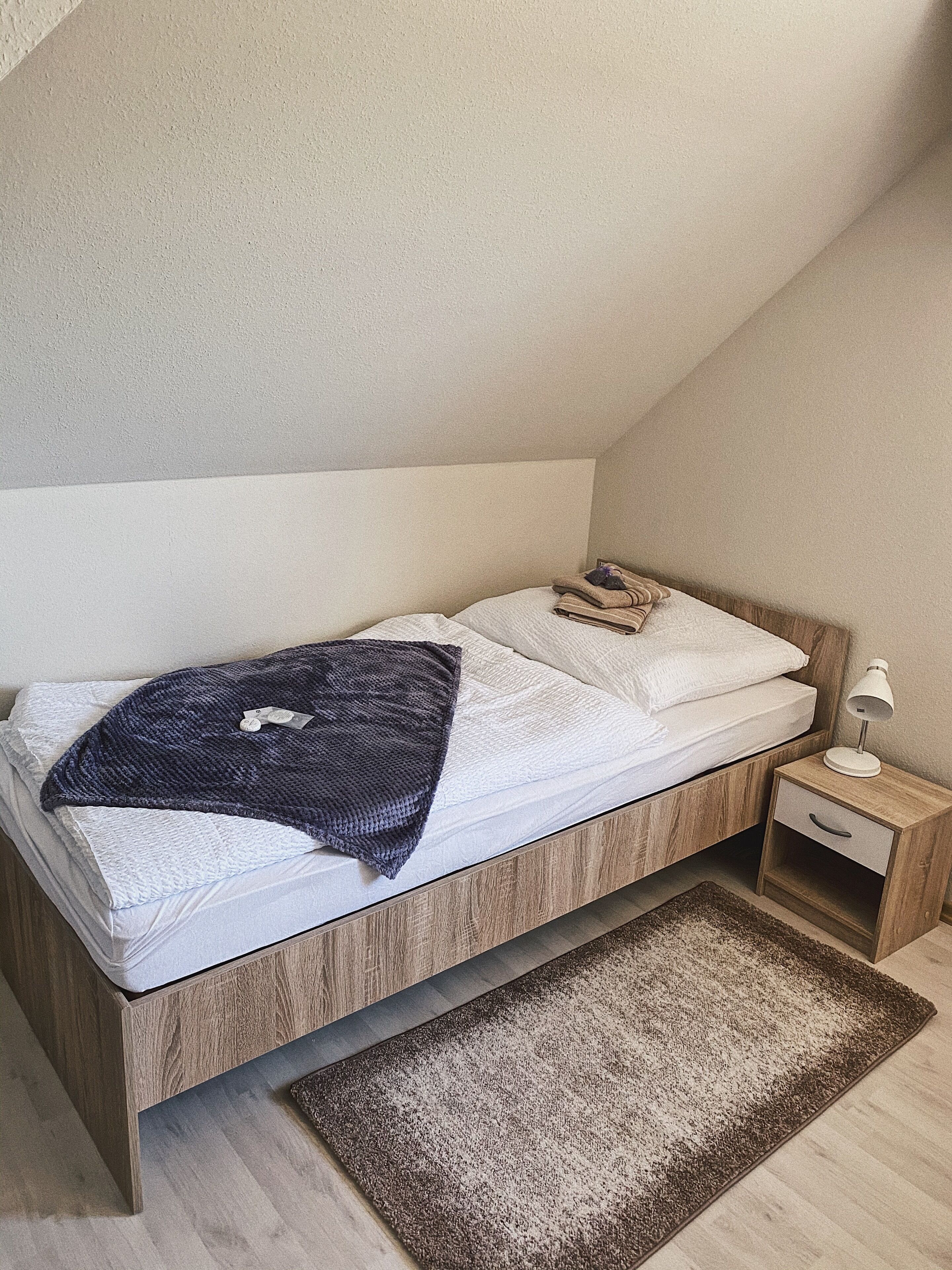 Un dormitorio con una cama de madera, ropa de cama blanca, un plaid oscuro, una mesita de noche con una lámpara y una alfombra marrón.