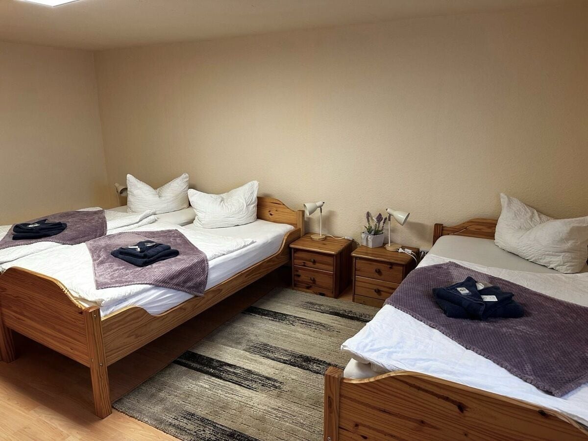 Ein Schlafzimmer mit zwei hölzernen Betten, jeweils mit weißer Matratze und Kissen, einem kleinen Nachttisch mit Lampe und einem Streifen Teppich auf dem Boden.