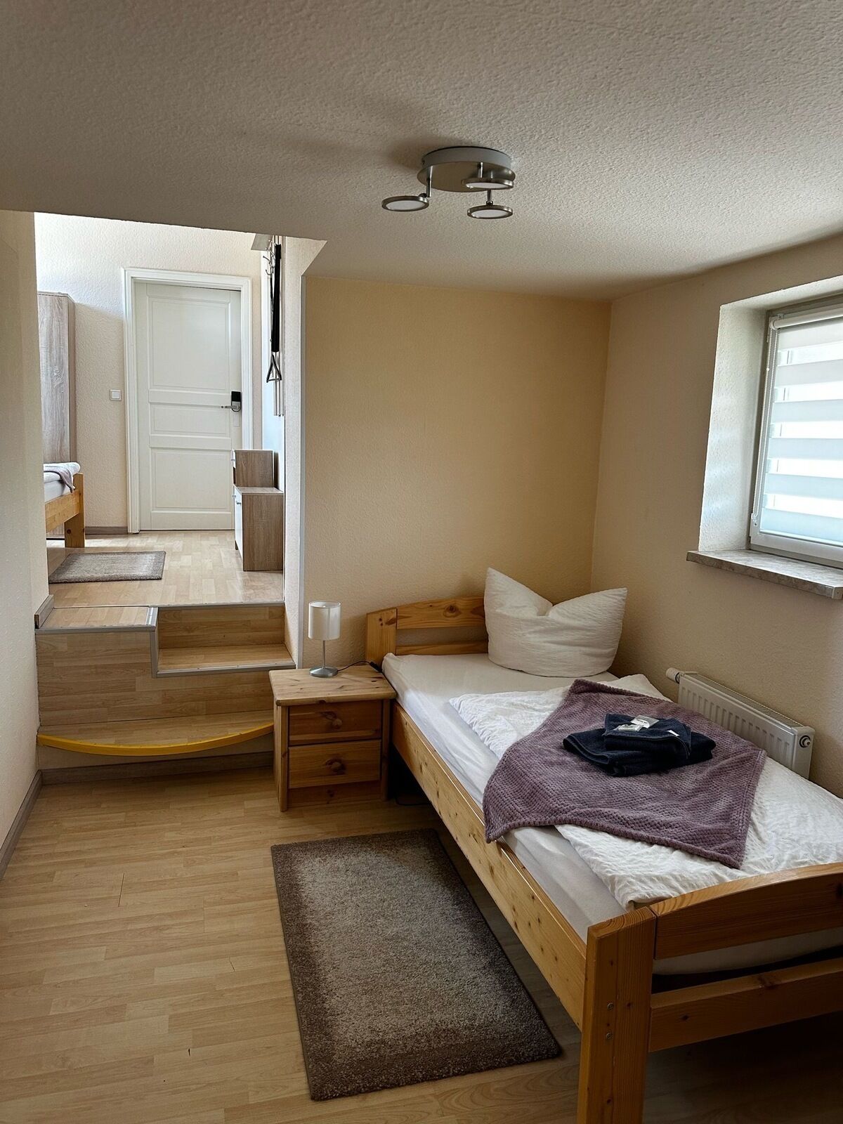 Un dormitorio pequeño y acogedor con una cama individual, una mesita de noche, un paralelo, una ventana con persianas y una puerta que da a otra habitación.
