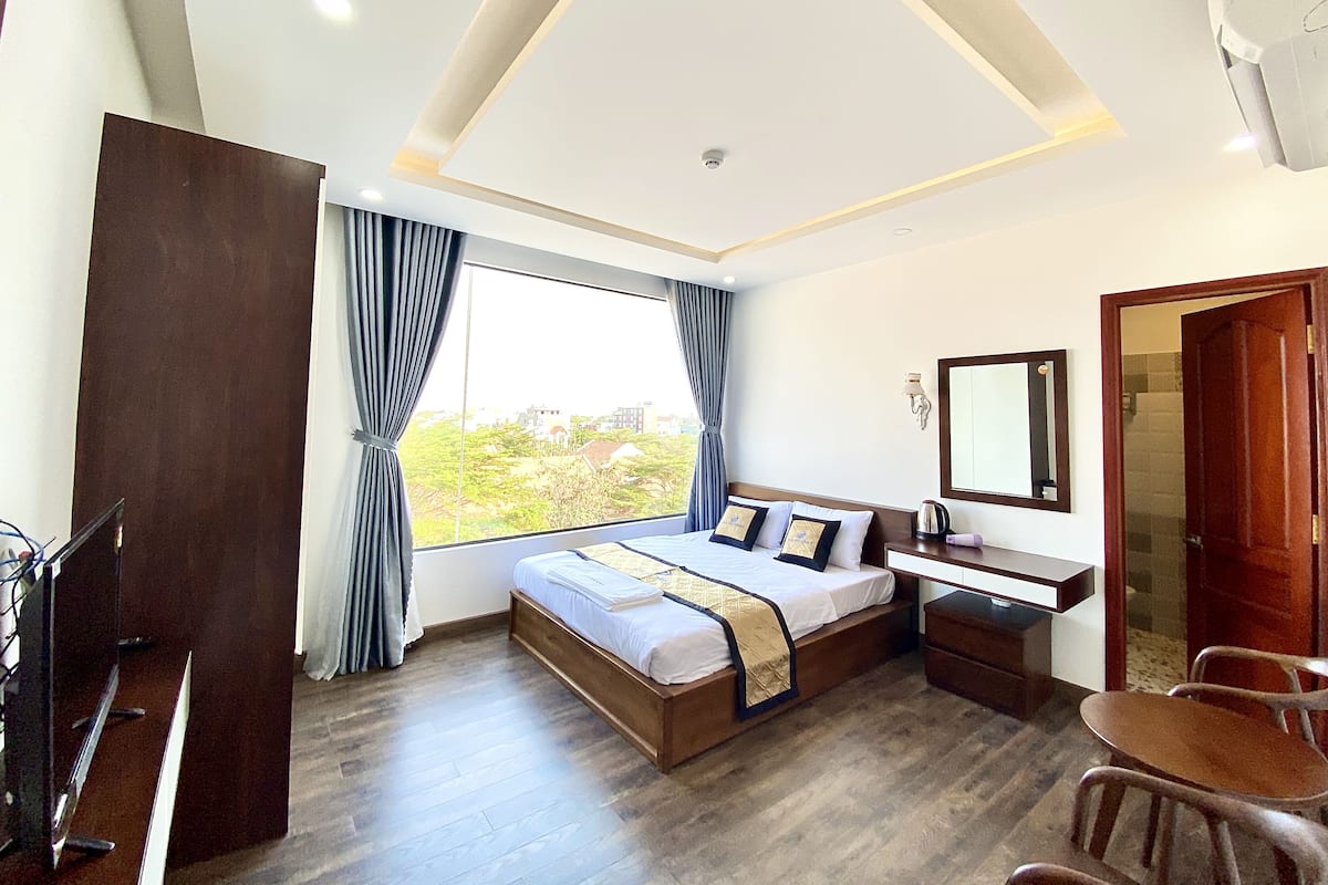 Quarto de hotel com uma cama grande, uma escrivaninha, uma cadeira e vista para o verde lá fora.