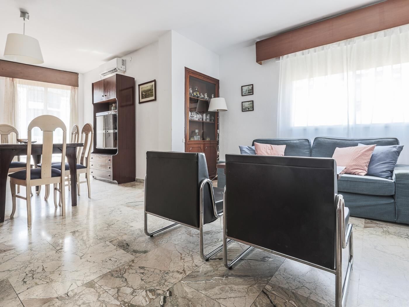 Una sala de estar con sofá, una mesa de comedor con sillas, un armario y un ventanal con cortinas.