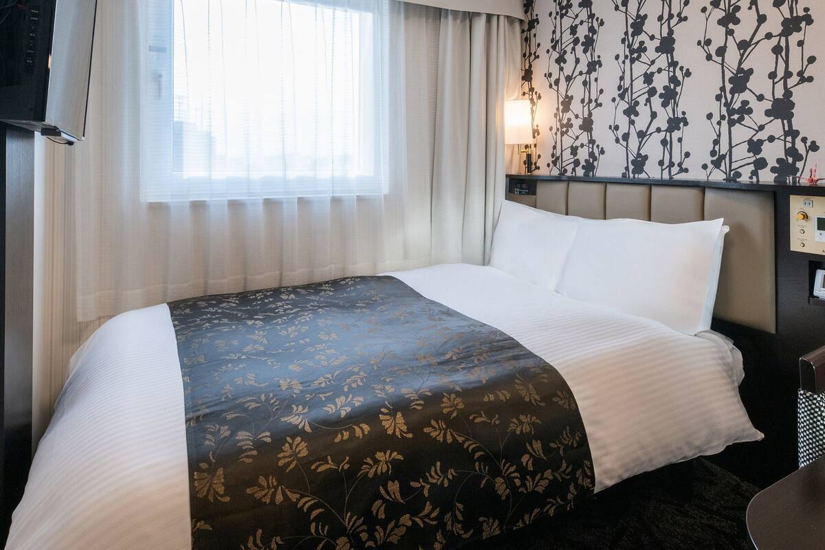A hotel room with a bed, a window with sheer curtains, a wall with a floral pattern, and a bedside table with a lamp.