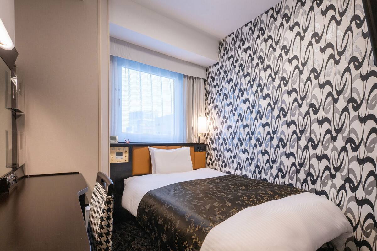 A hotel room with a bed, a bedside table, a lamp, a window with curtains, and a patterned wall.