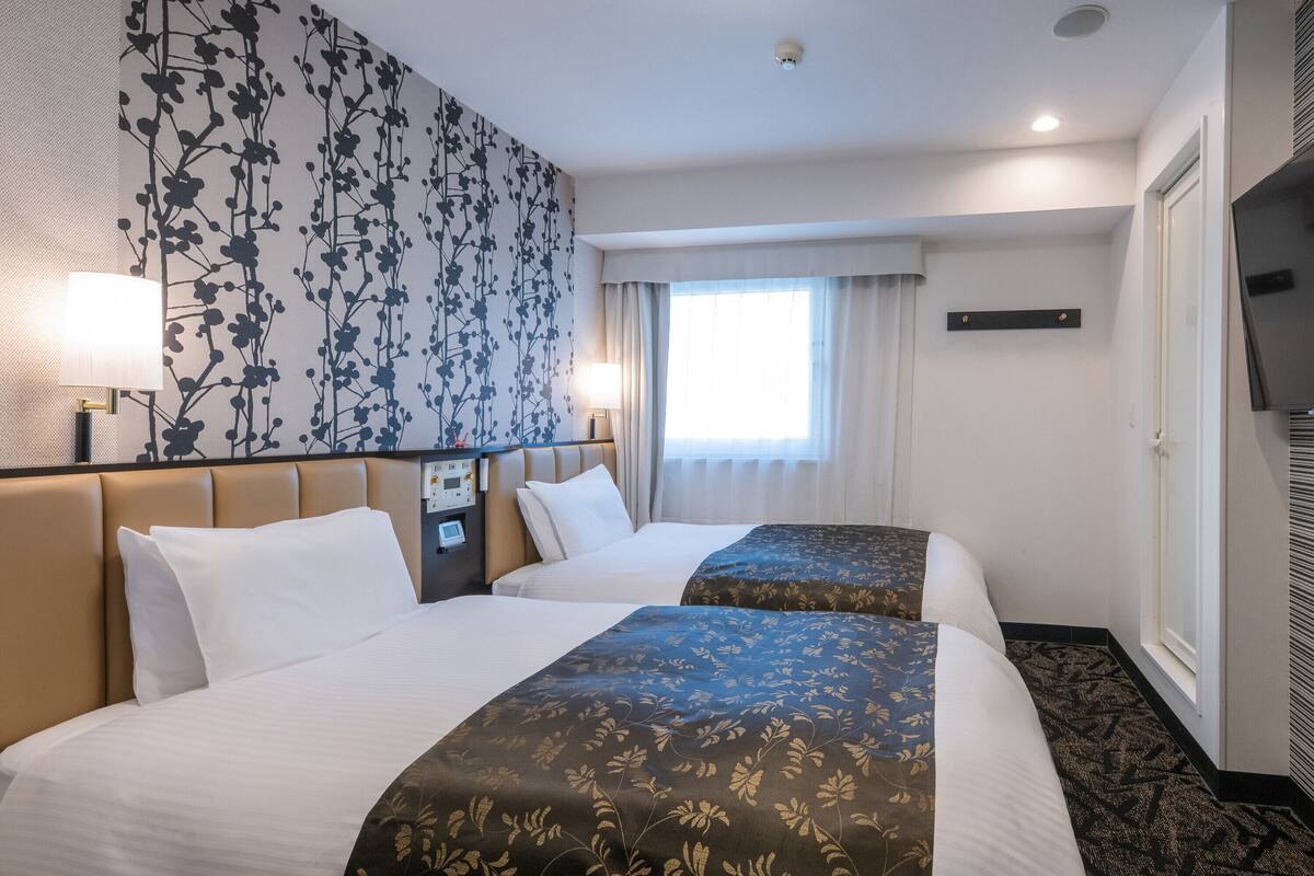 A hotel room with two beds, a large window with curtains, a flat-screen TV mounted on the wall, and a control panel on the nightstand.