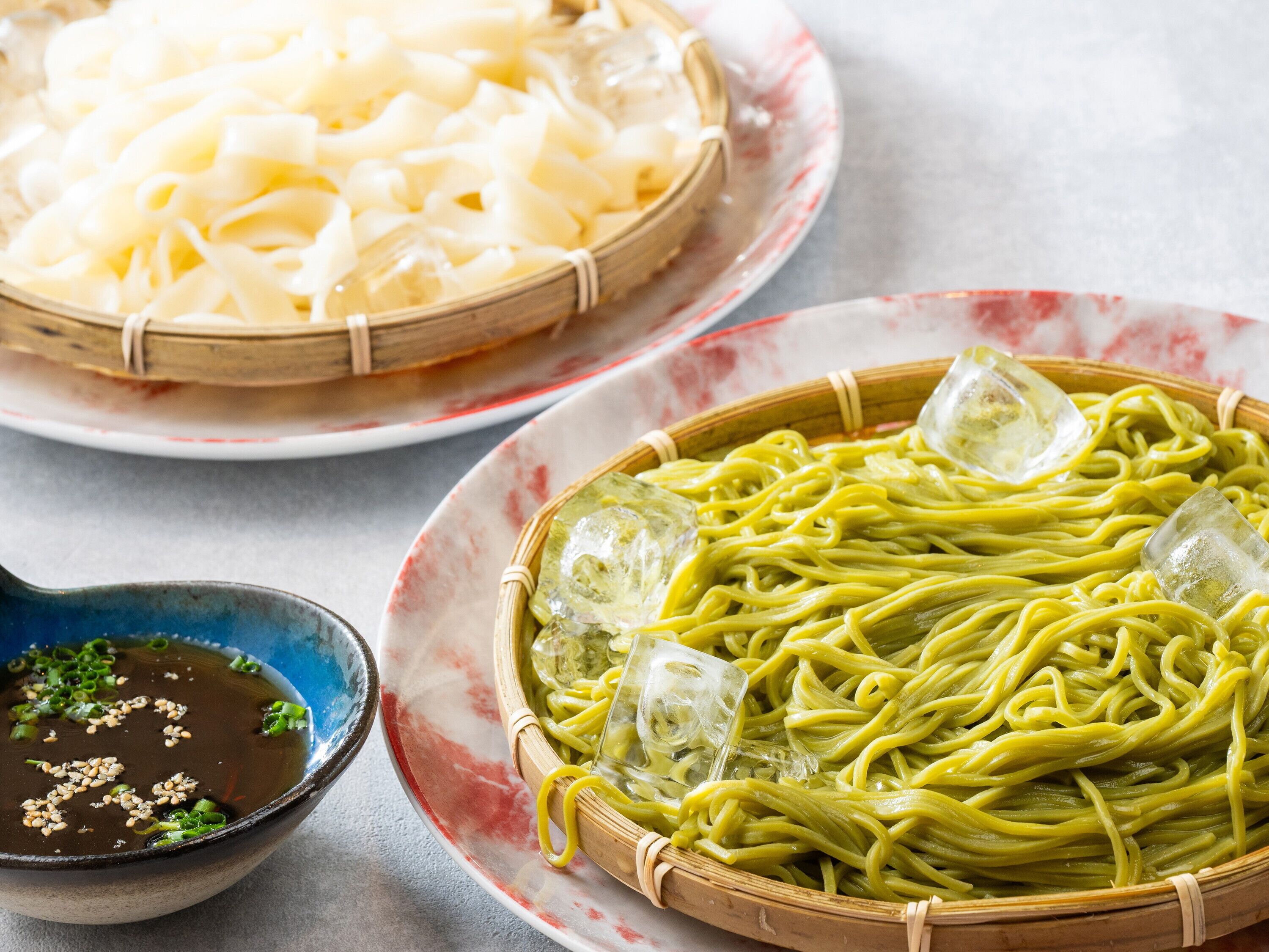 A bowl of soup with green onions and sesame seeds, a plate of noodles, and a plate of green noodles with ice cubes.
