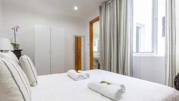Ein Hotelzimmer mit einem Bett, zwei Kissen, einem Nachttisch mit Lampe, einer Topfpflanze und einem Fenster mit gestreifter Vorhang.