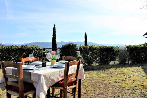 An outdoor dining table set with plates, glasses, and a bottle, surrounded by wooden chairs, with a view of distant trees and hills. - Villa Bagnano (Certaldo)