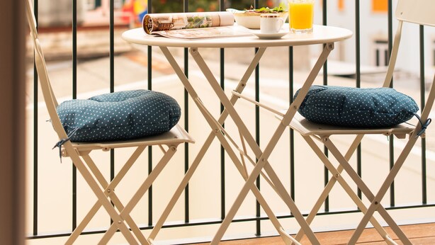 A balcony with a table and chairs, a glass of orange juice, and a view of a cityscape with buildings and a tower. - Apartamentos Ferreiros (Funchal)