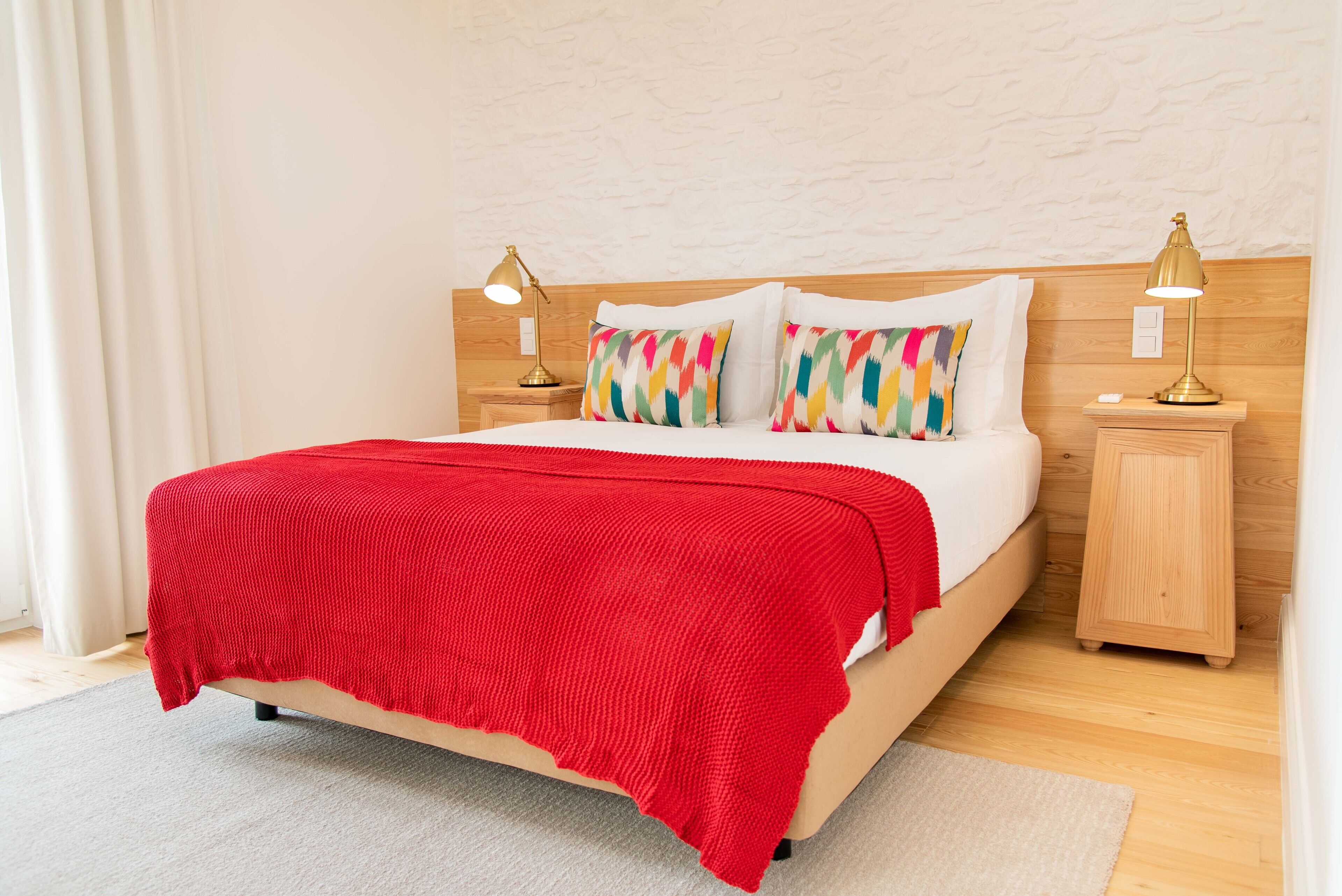 A neatly made bed with a red blanket, two bedside lamps, and colorful pillows.