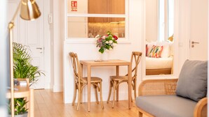 A room with a wooden table, two chairs, a sofa, and a window with a view of a kitchen. - Apartamentos Ferreiros (Funchal)
