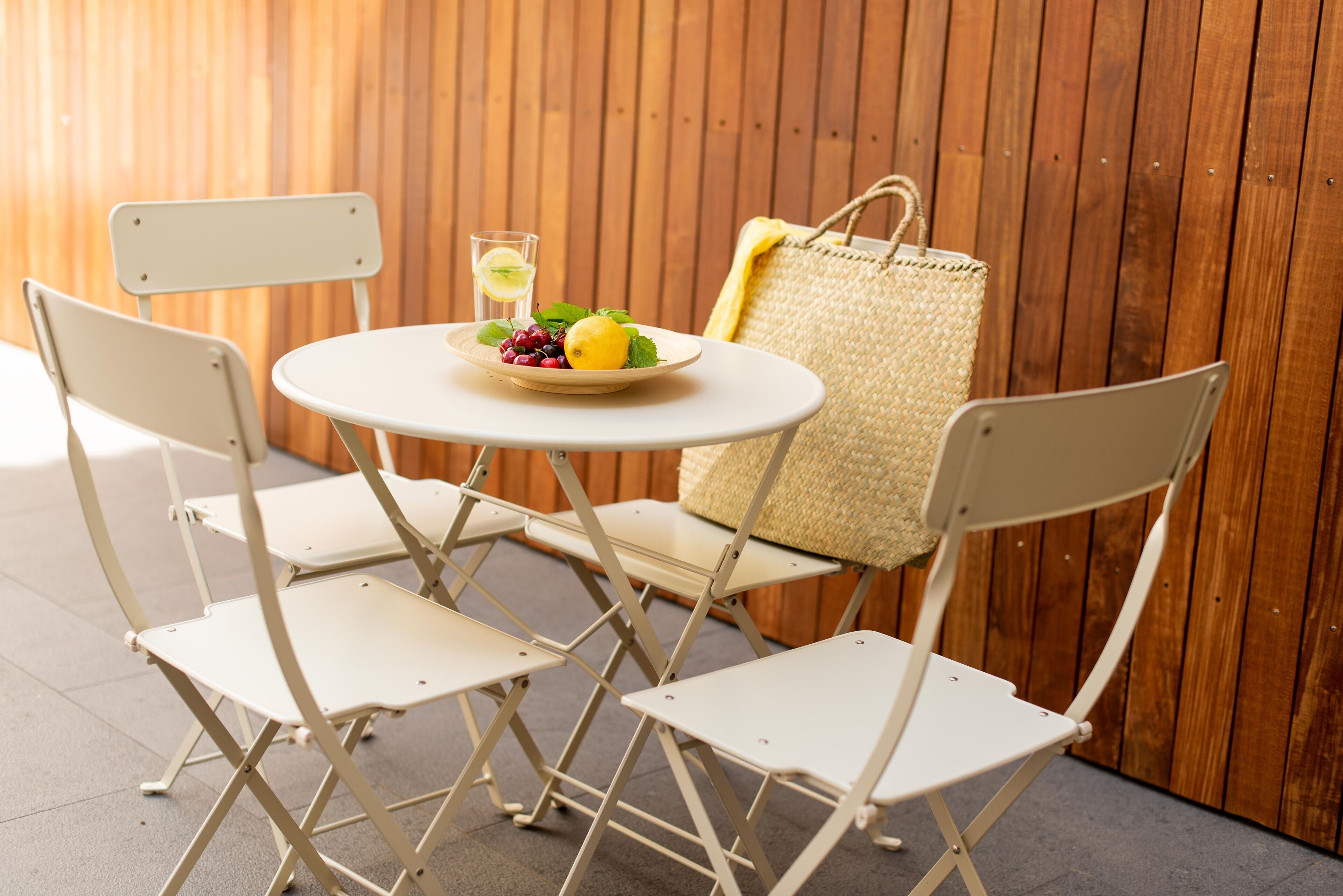 A small outdoor seating area with a round table, two white chairs, a wicker bag, and a plate of fruit and a glass of drink.