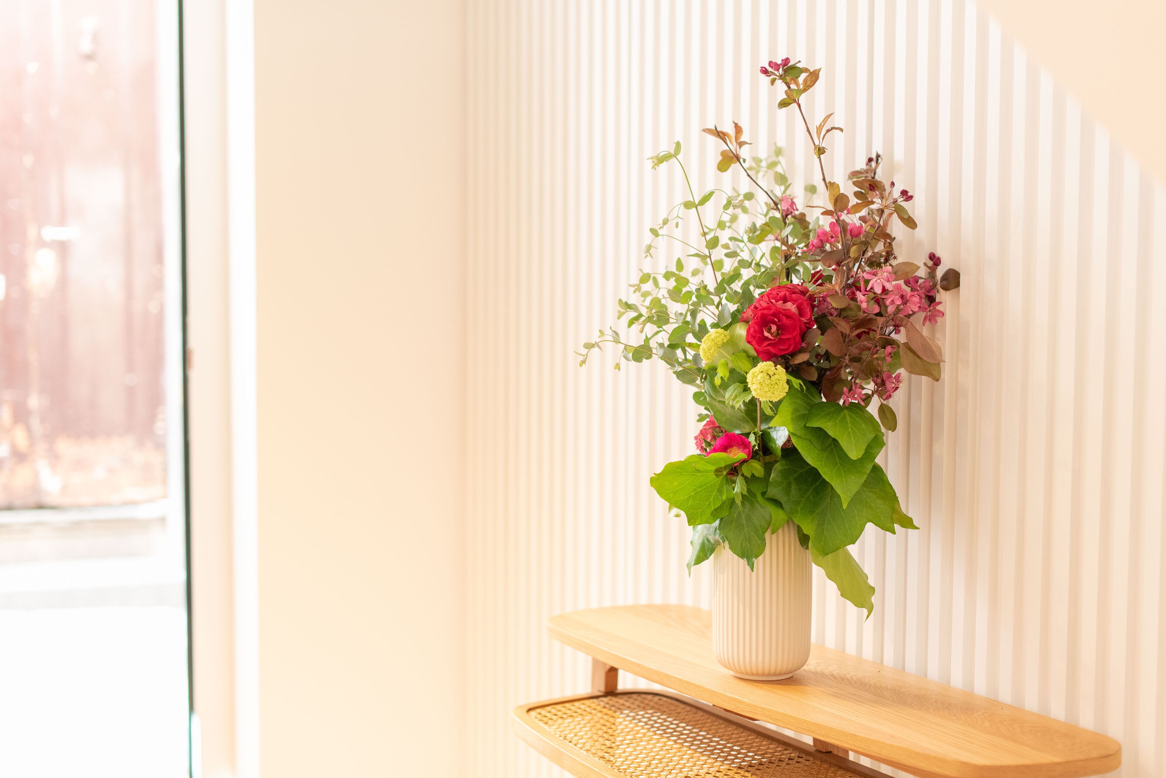 A vase with flowers on a wooden shelf against a wall with vertical blinds.