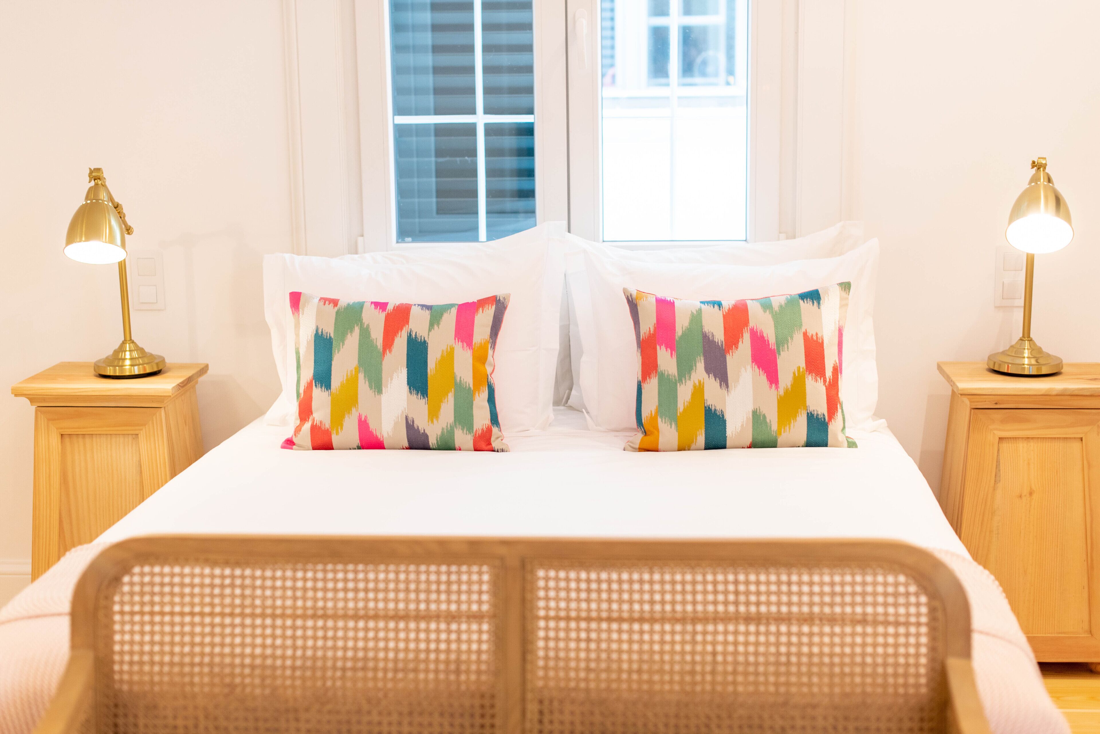 A neatly made bed with colorful pillows, two bedside lamps, and a window with blinds.
