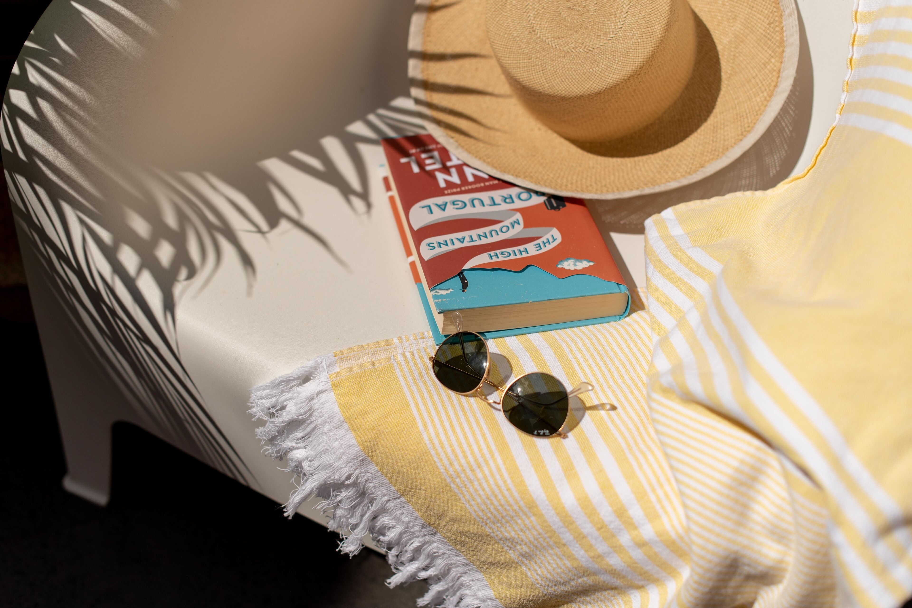 A beach chair with a straw hat, a book titled 'The Mountainous Road', and sunglasses on a striped towel.