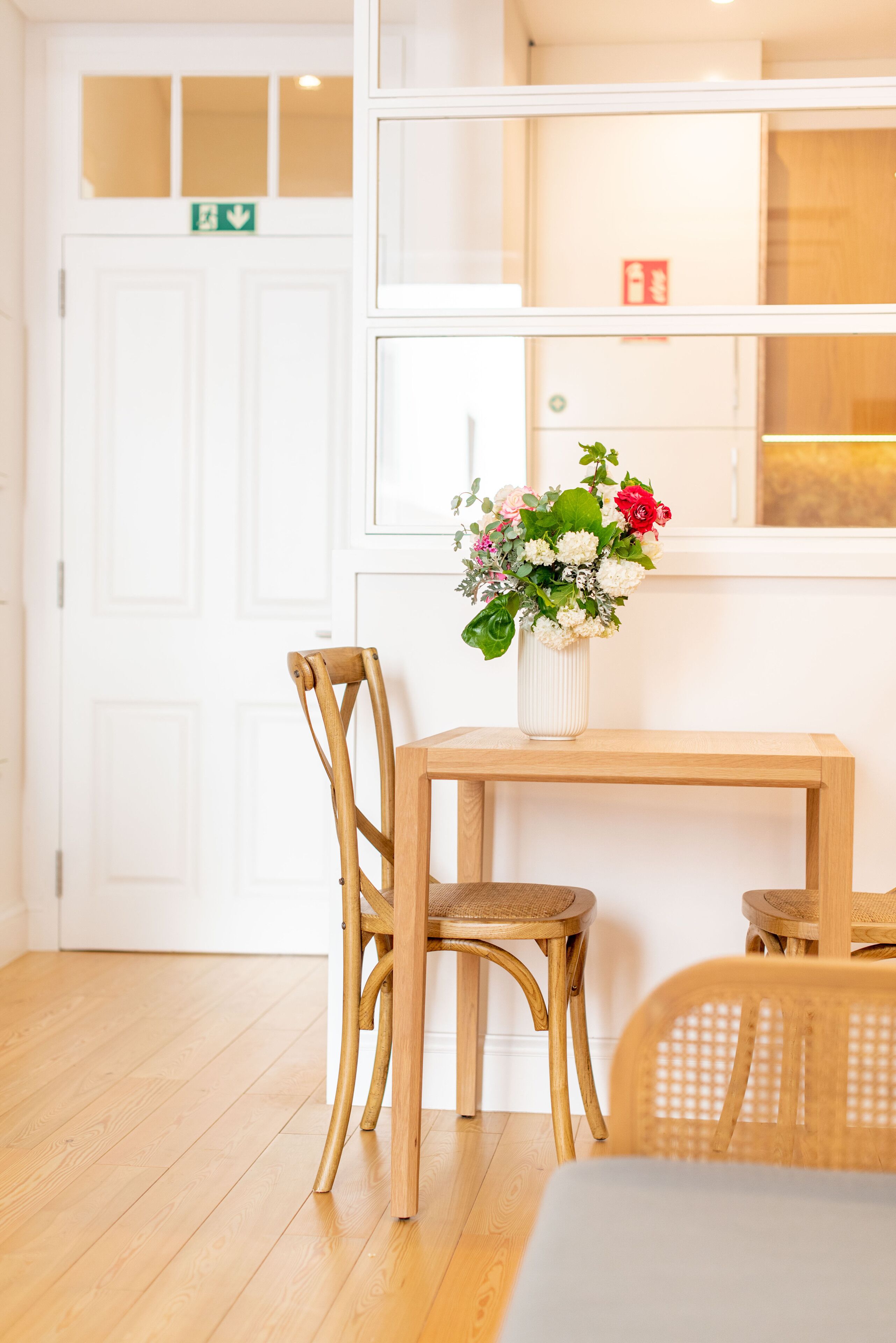 Ein Holztisch mit einem Blumenstrauß, zwei Korbstühle und eine weiße Tür mit einem Hinweisschild für den Ausgang.