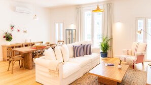 A bright living room with a white sofa, wooden coffee table, and a dining area with a wooden table and chairs. - Apartamentos Ferreiros (Funchal)