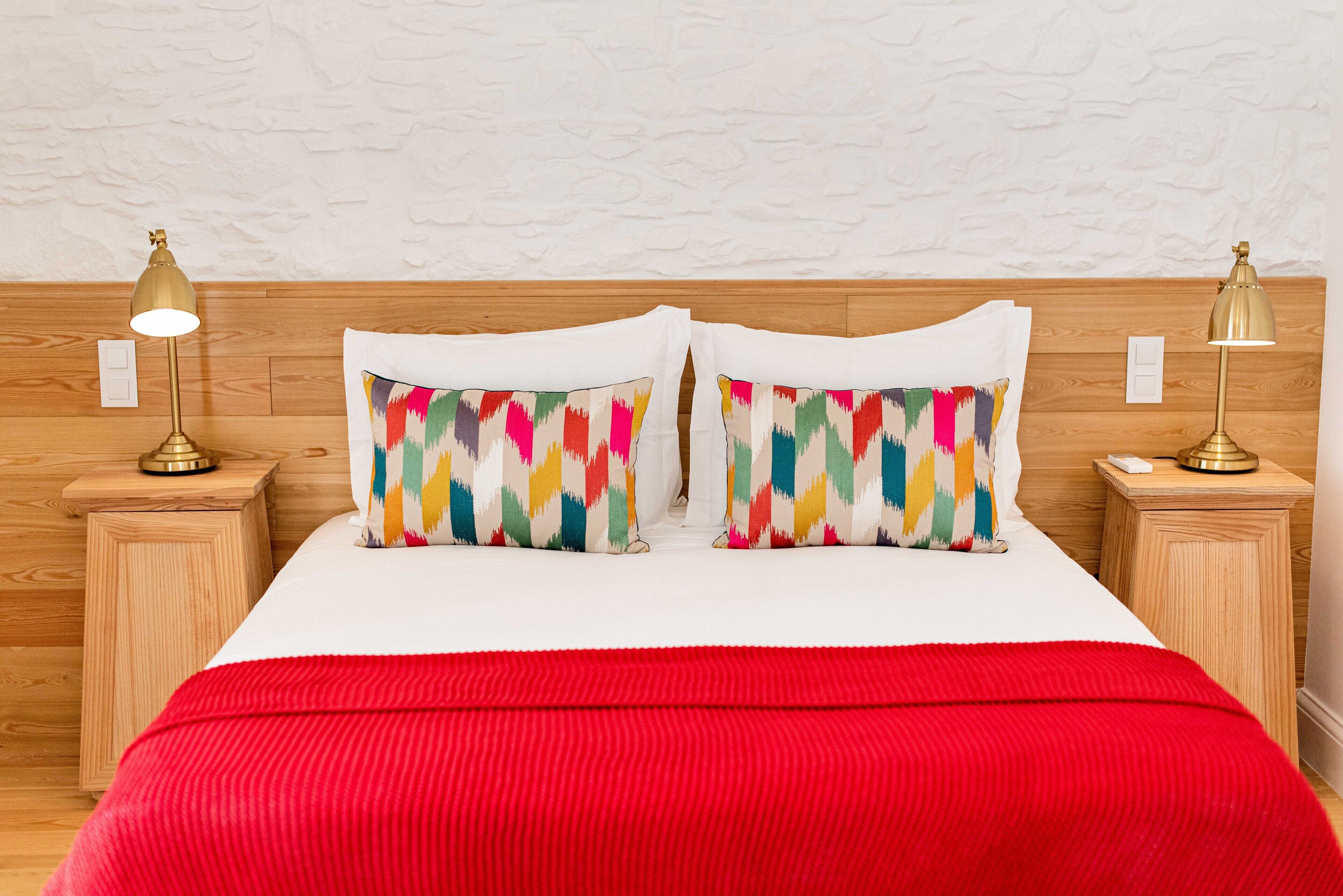Un letto con una coperta rossa, due cuscini colorati e due lampade da comodino su mensole in legno.