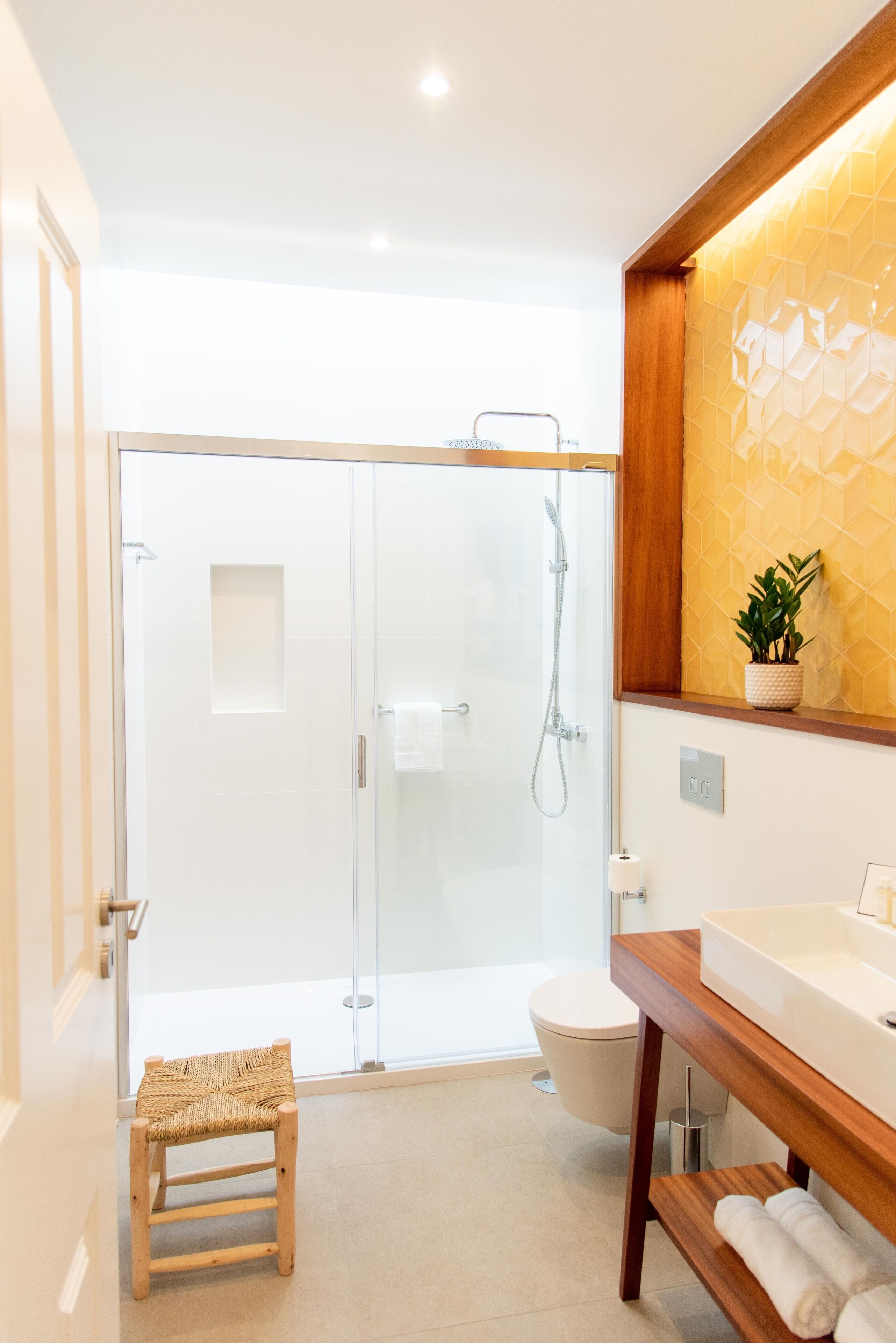 Une salle de bain moderne avec une douche en verre, un banc en bois, un lavabo blanc et une plante en pot.