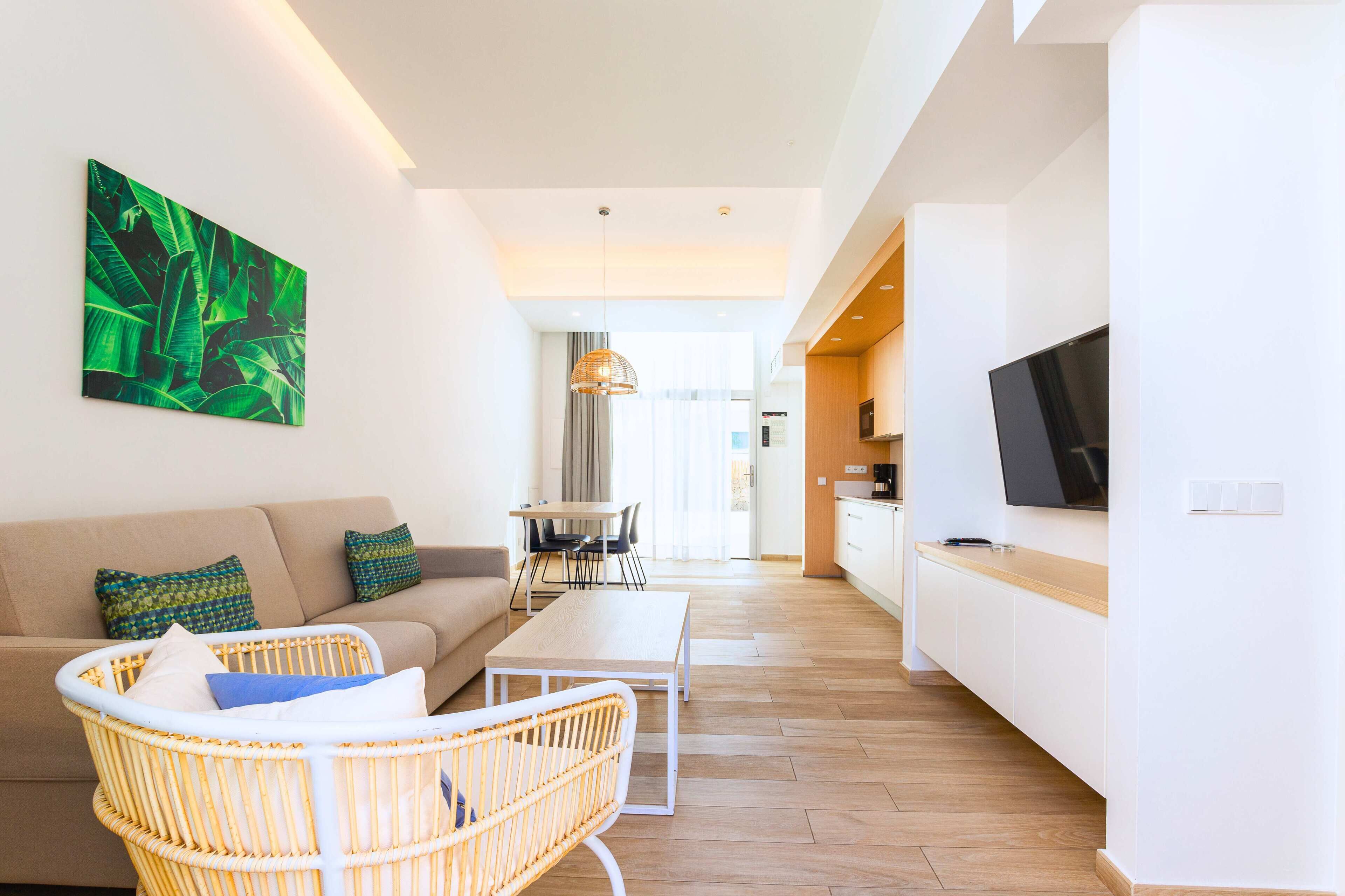 A modern living room with a sofa, wicker chairs, a coffee table, and a dining area with a table and chairs. A kitchen with a TV and cabinets is visible in the background.