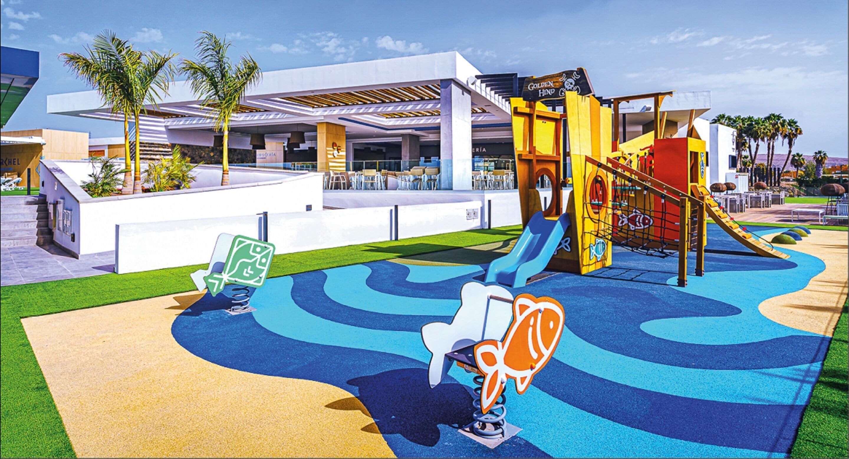 A playground with slides and swings, surrounded by a modern building with a flat roof and palm trees.