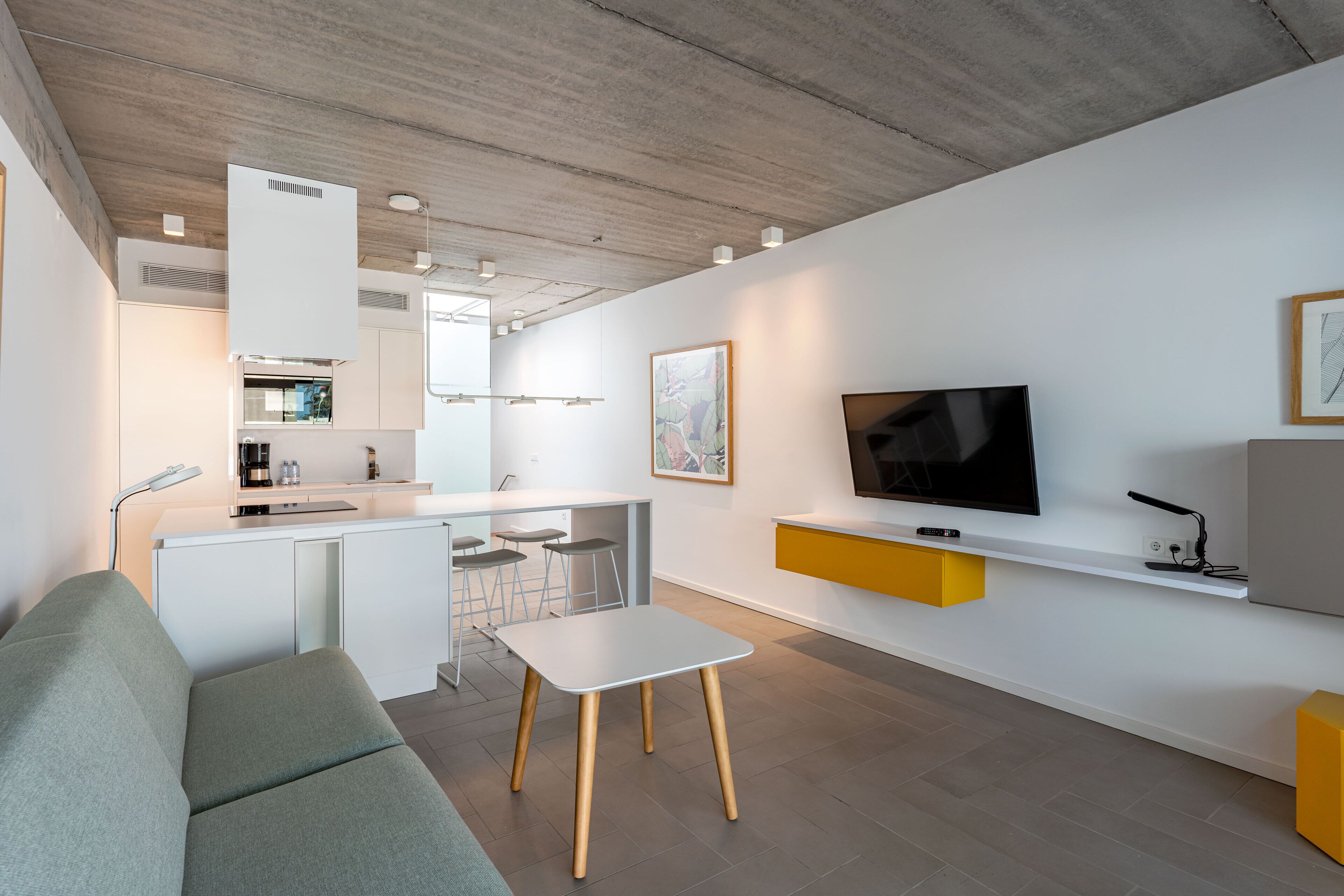 Un espacio habitable moderno con sofá, una mesita, una zona de cocina y un televisor montado en la pared.