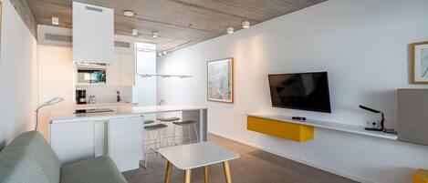 Un espacio habitable moderno con sofá, una mesita, una zona de cocina y un televisor montado en la pared.