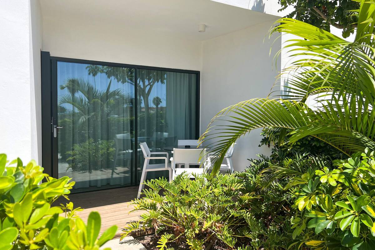 Eine moderne Außenpatio mit Glastür zu einem Balkon, umgeben von üppiger Vegetation und weißen Gartenmöbeln.