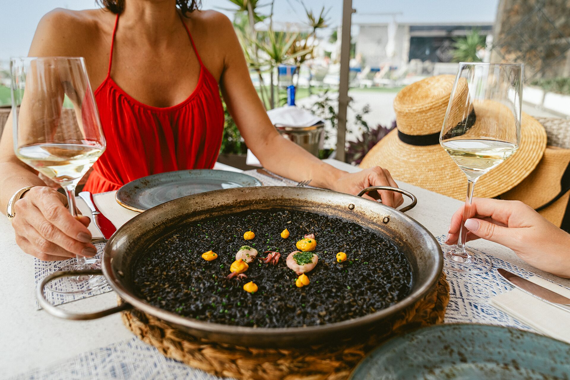 Une table à manger sur laquelle se trouve un grand plat de risotto noir, garni d’herbes et de crevettes, et deux personnes tenant des verres de vin.