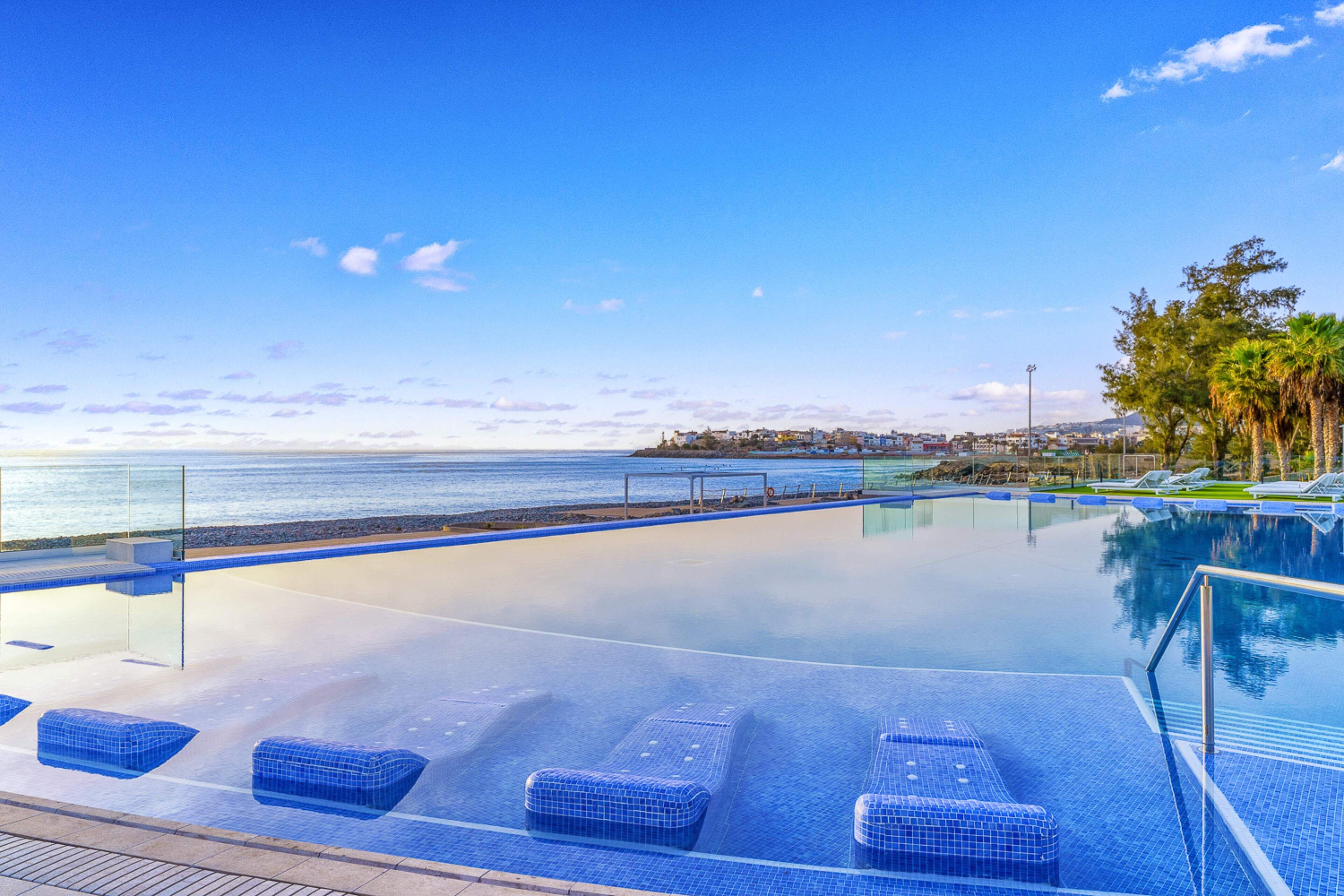 Un cielo azul despejado, mar tranquilo y una piscina con sillones de descanso.