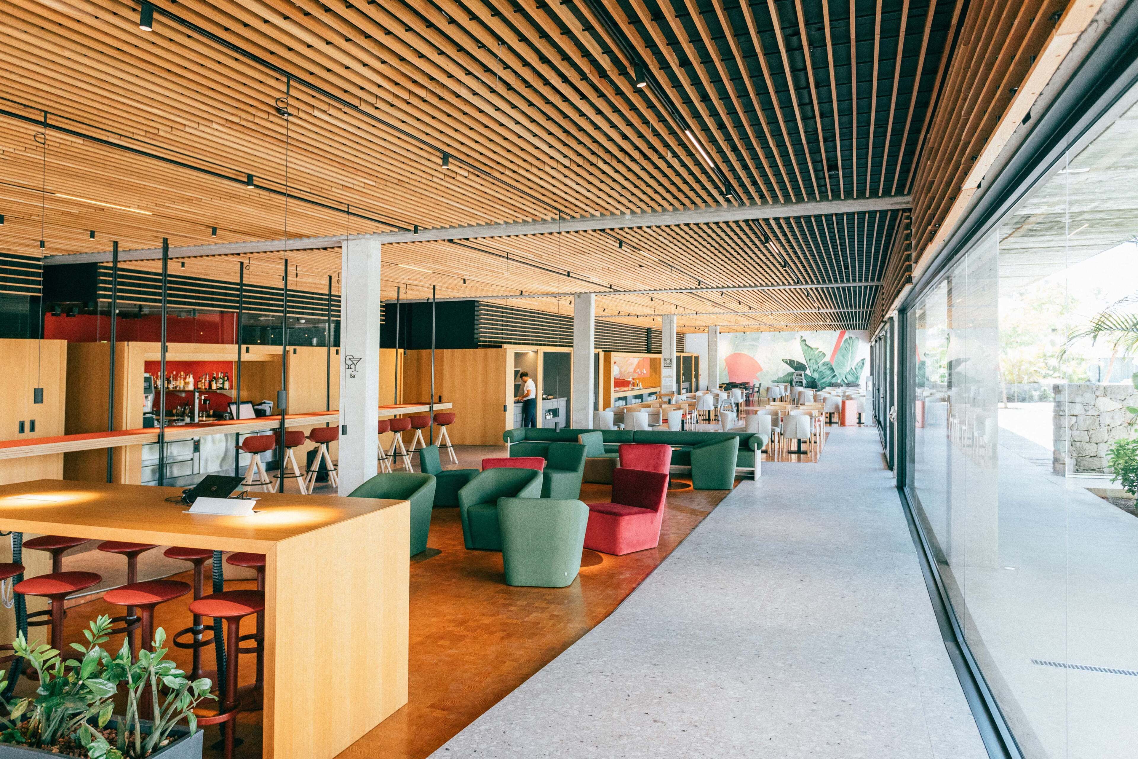 Un espacio interior moderno con techos de listones de madera, áreas de estar en tonos verde y rojo, y una barra con taburetes.