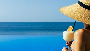Une personne tient un cocktail décoré d’une tranche d’orange, porte un chapeau de paille et se trouve près d’une piscine à l’eau cristalline, avec une vue sur l’océan.