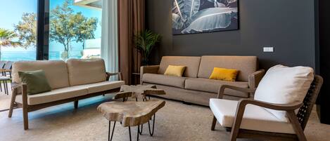 A modern living room with a sofa set, a wooden coffee table, and a chair. There is a large window with a view of the sea and a plant in the corner.