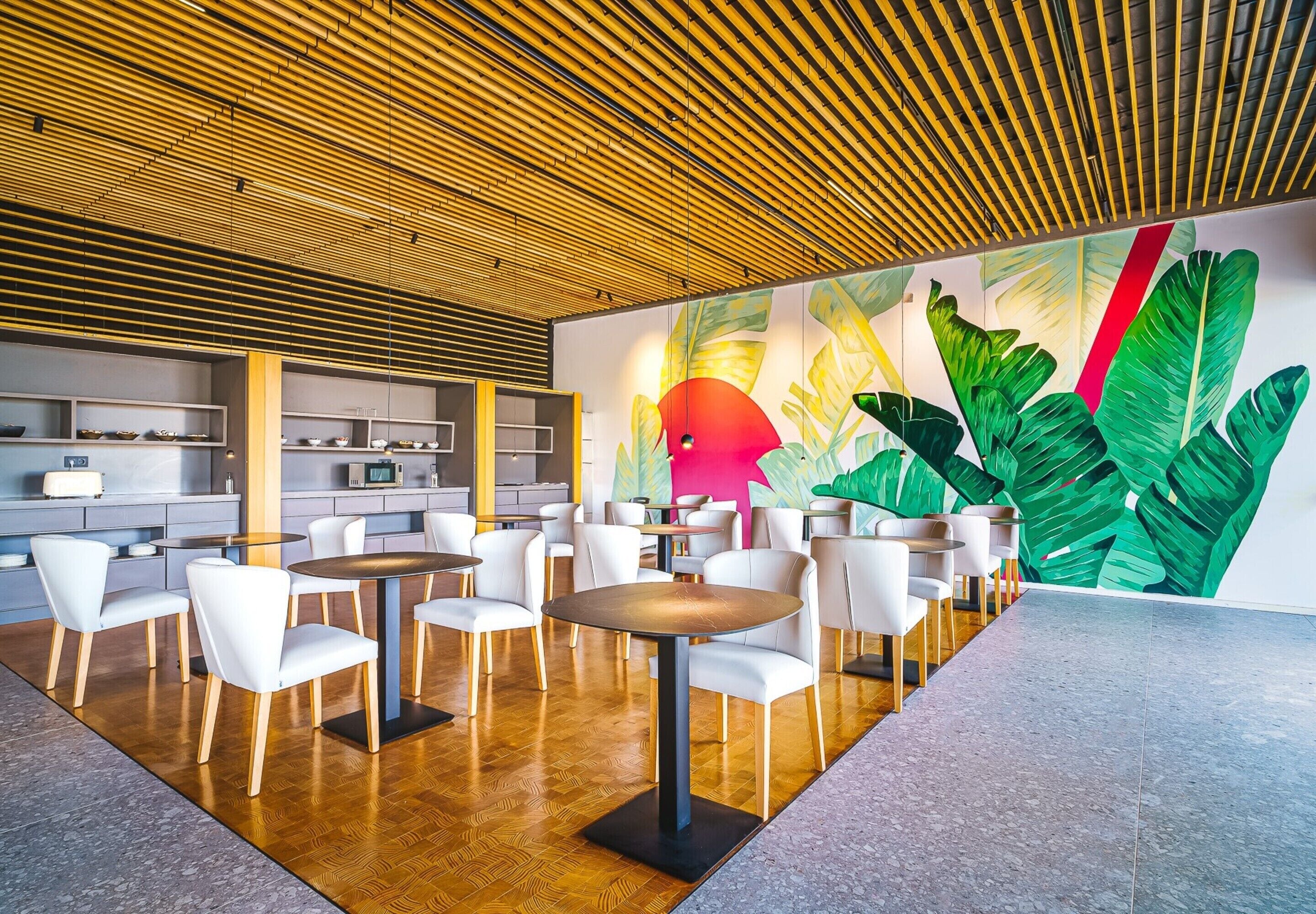 Un interior moderno con mesas redondas y sillas blancas, techo de madera y un mural vibrante con hojas grandes y un círculo rojo.