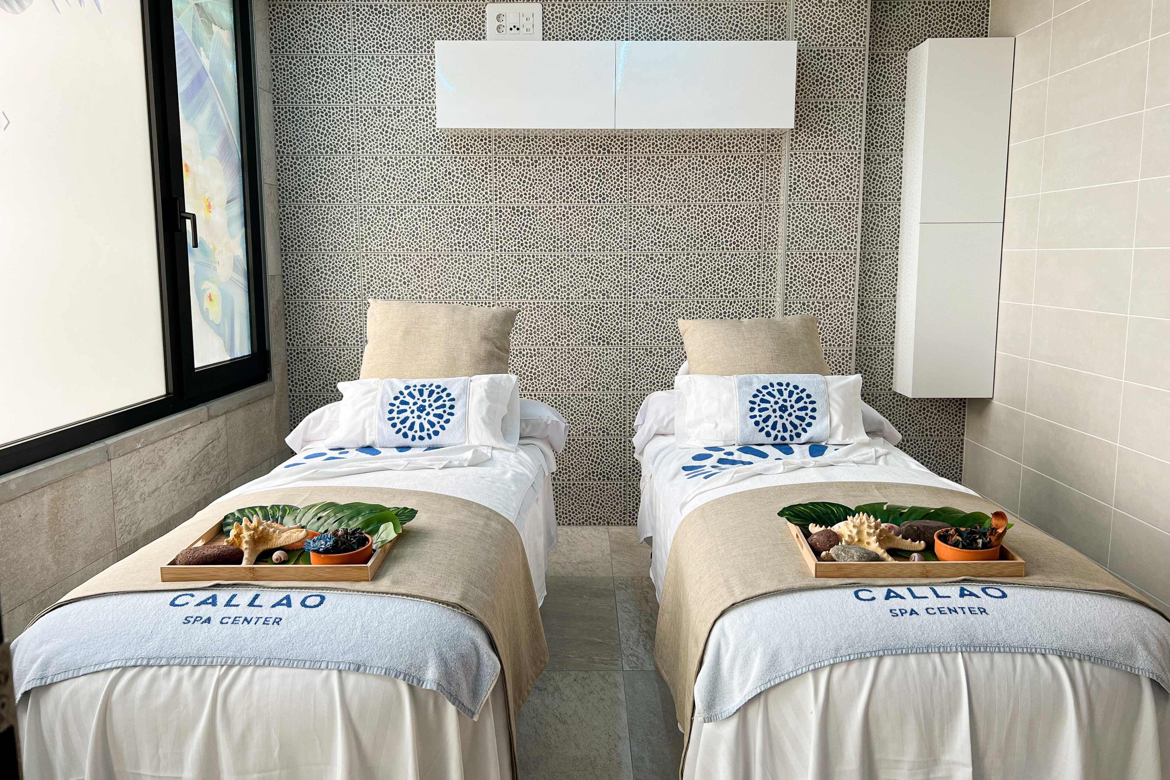 A hotel room with two beds, each with a tray of fruits and a towel with "CALLAO SPA CENTER" printed on it.