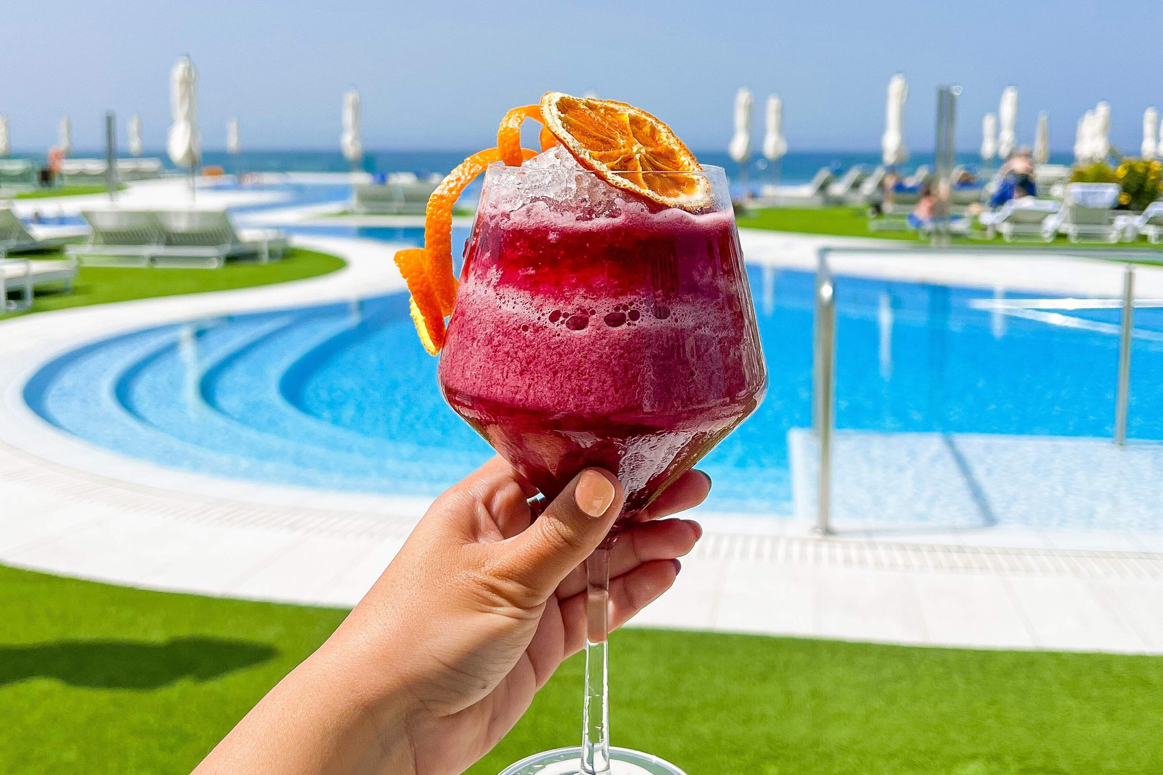 Una mano sosteniendo un vaso con una bebida roja y una rodaja de naranja, con una piscina y sillas de descanso al fondo.