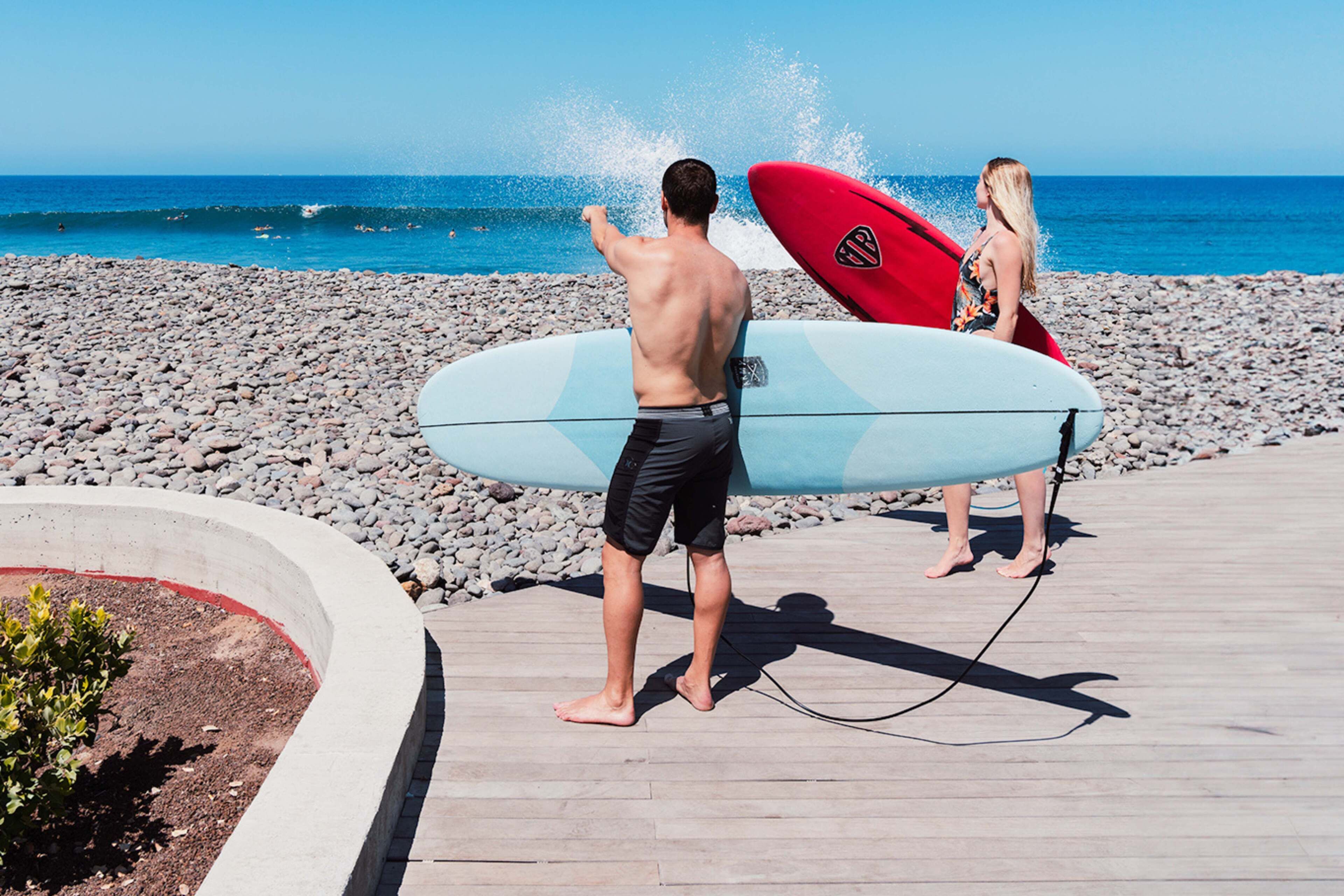 A wooden boardwalk leading to a beach with two people holding surfboards.