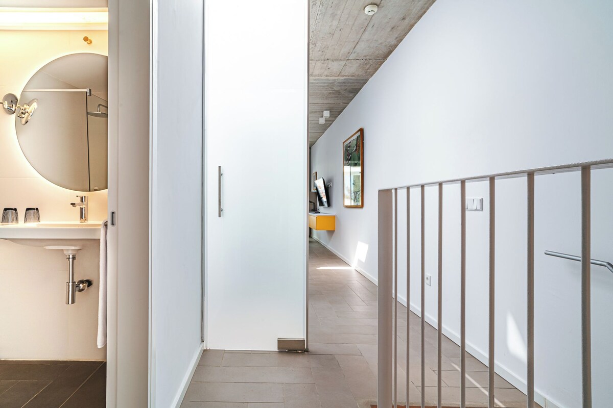 Un pasillo con pared blanca, un espejo redondo, una puerta blanca y una barandilla de metal.