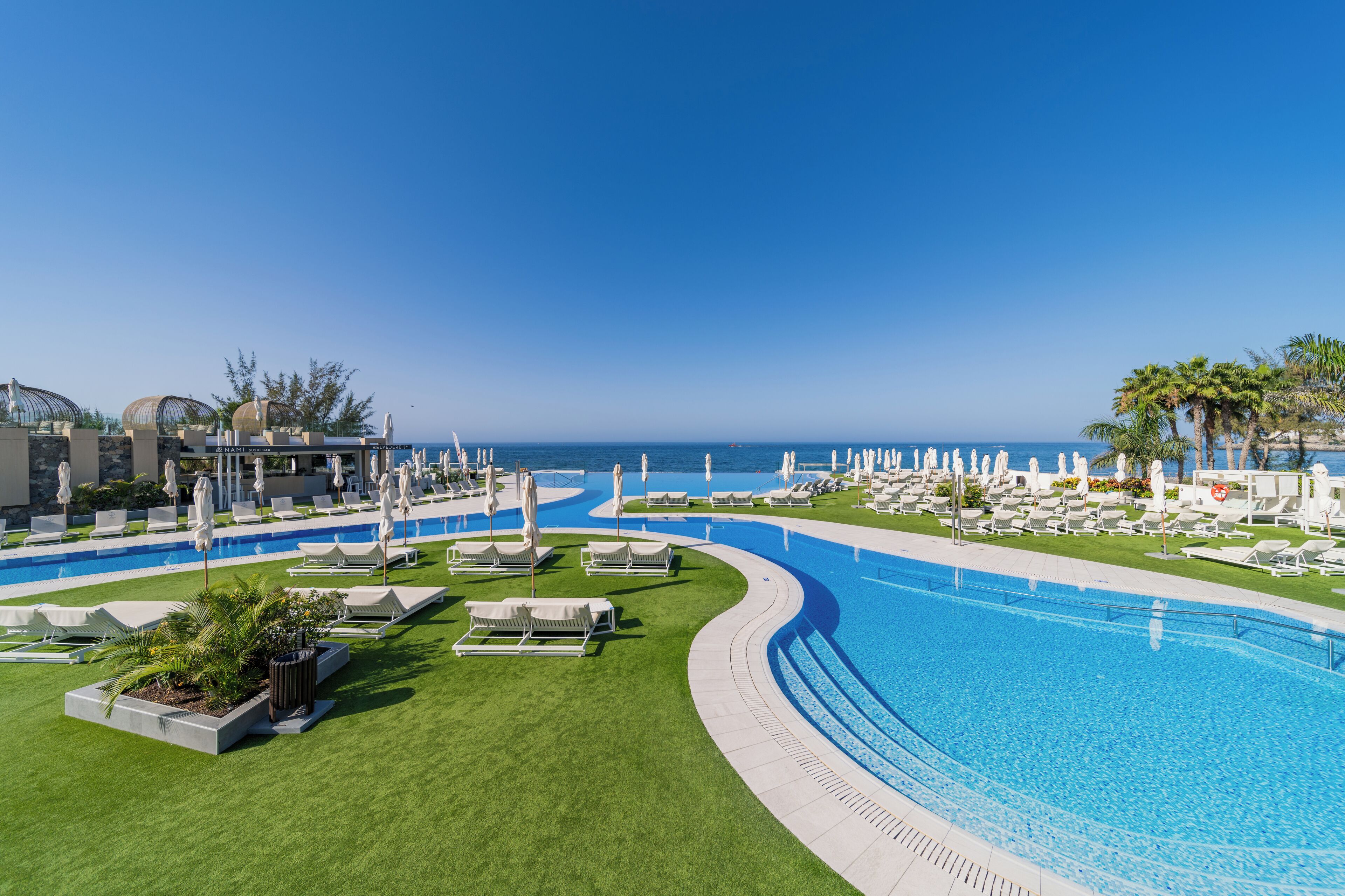 Área de piscina de resort com piscina sinuosa, espreguiçadeiras e guarda-sóis, com vista para o oceano.