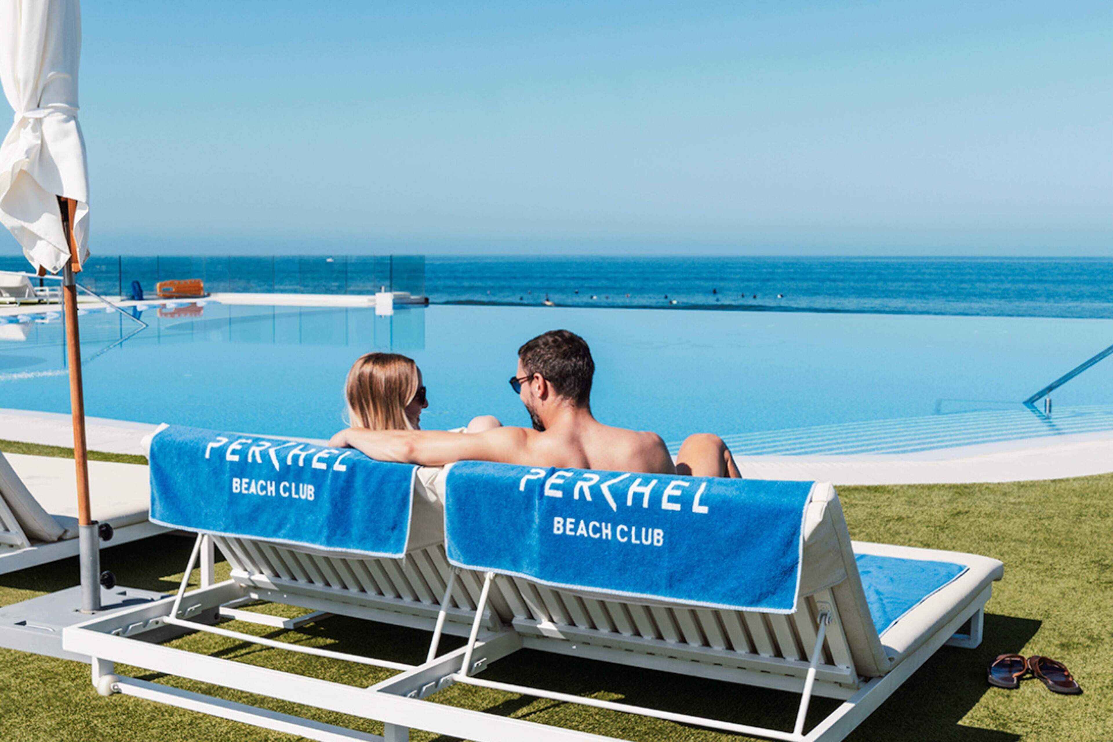 Two lounge chairs with blue towels labeled "Perhel Beach Club" facing a pool and ocean view.