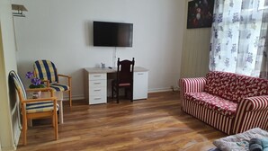 Ein Zimmer mit Holzboden, einem Schreibtisch mit Stuhl, einem an der Wand befestigten Fernseher, einem Sofa und einem Fenster mit Vorhängen.