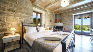 A bedroom with stone walls, a wooden ceiling, a bed with white bedding, a nightstand with a lamp, and a view of greenery through a window. - Asion Suite Eros with Private Pool (Kato Asites)