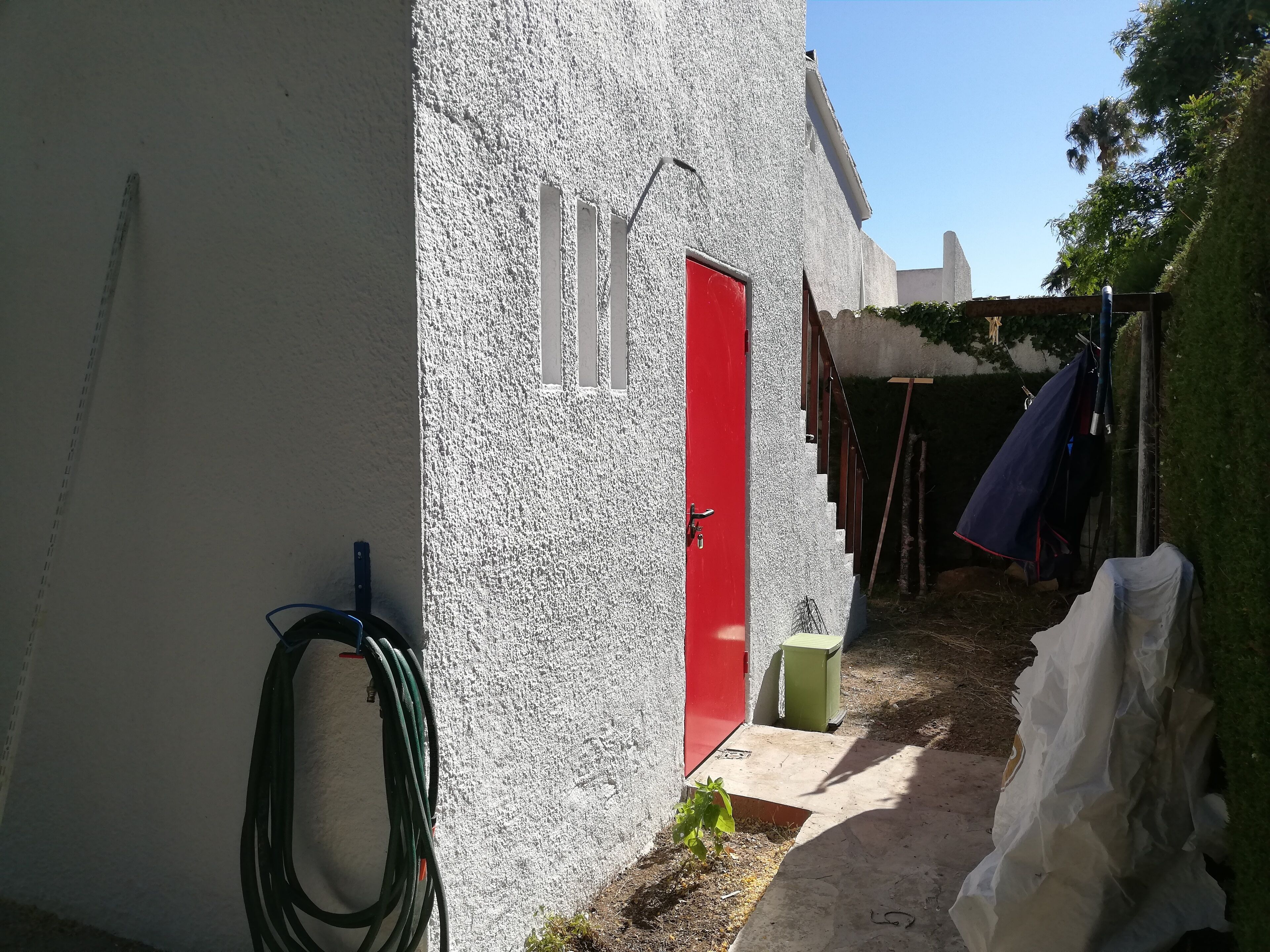 A narrow outdoor pathway leading to a building with a red door and a white exterior.