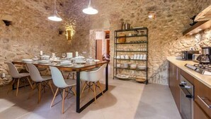 Un intérieur rustique en pierre, avec une table à manger dressée pour quatre personnes, un coin cuisine équipé d’appareils modernes et une étagère remplie de vaisselle.