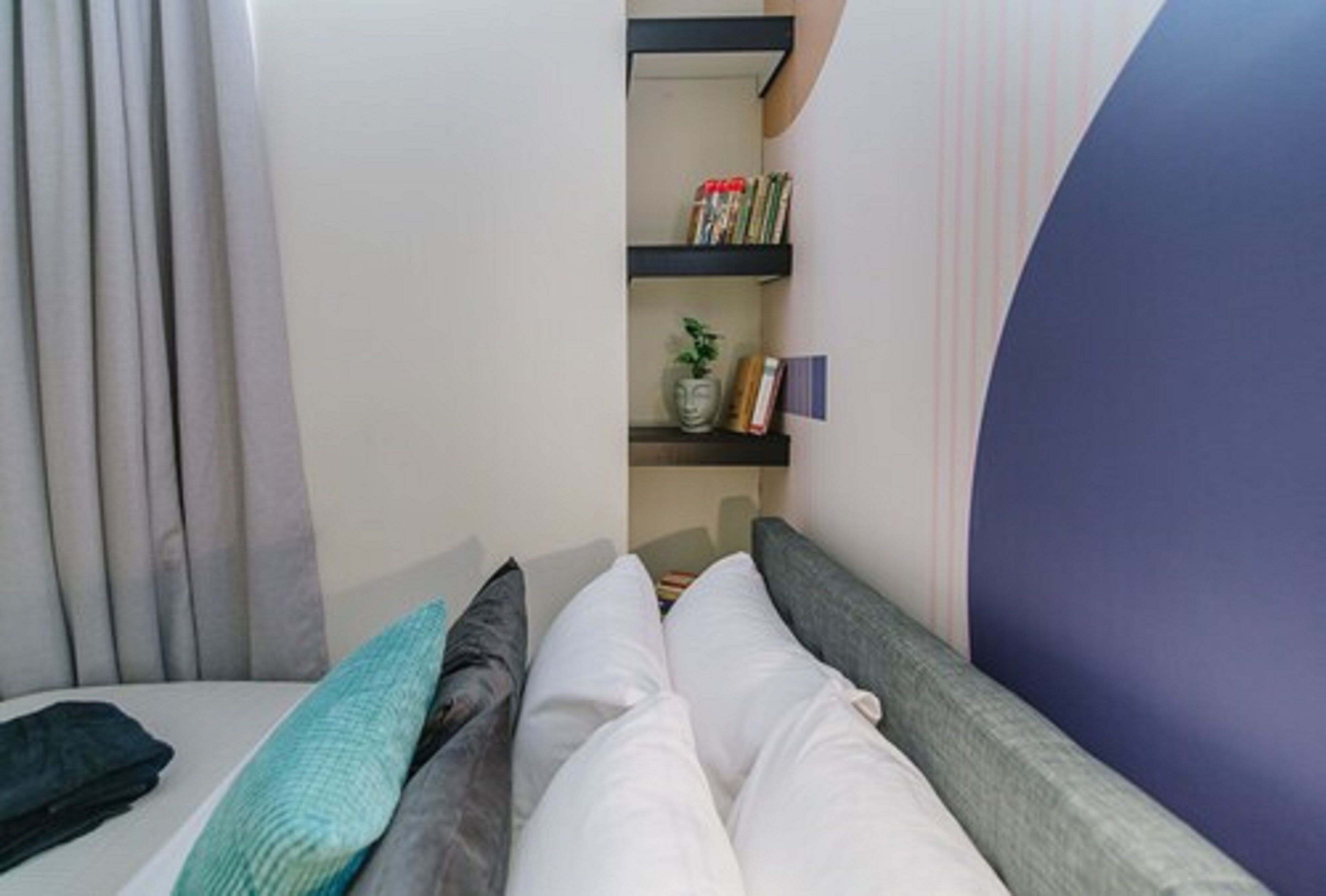 A bedroom with a bed, pillows, a nightstand, a shelf with books and a vase, and a window with curtains.